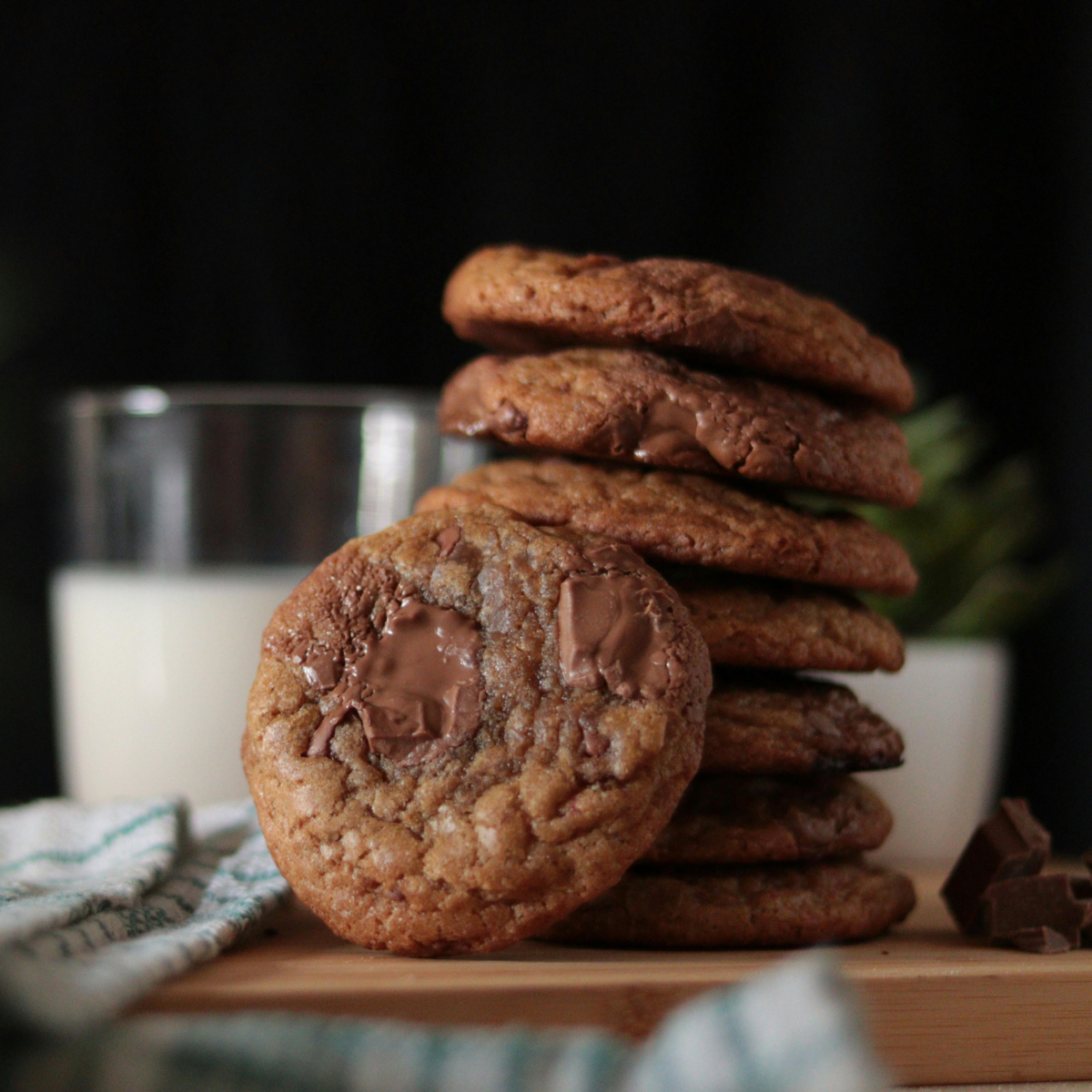 Stack of chocolate chip cookies with one cookie in front showing melted chocolate, in front of a glass of milk.