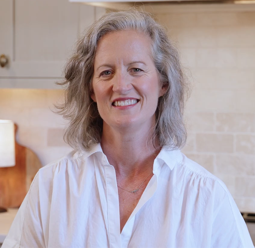 A smiling woman with shoulder-length gray hair wearing a white button-up shirt in a kitchen setting. The pastry chef behind Mookie.