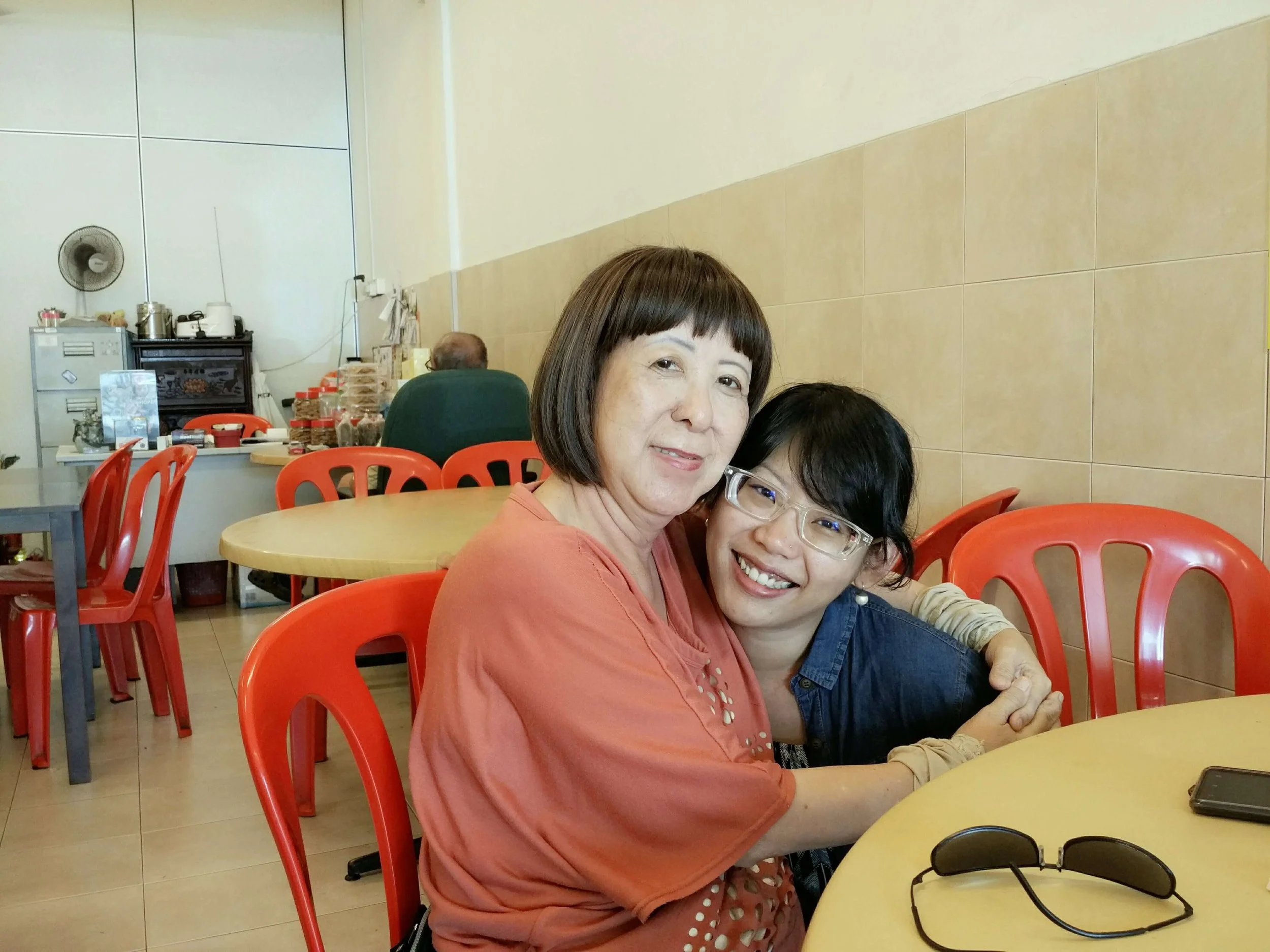 Two women sitting together at a round table in a restaurant, smiling at the camera, with red chairs and a beige wall in the background.