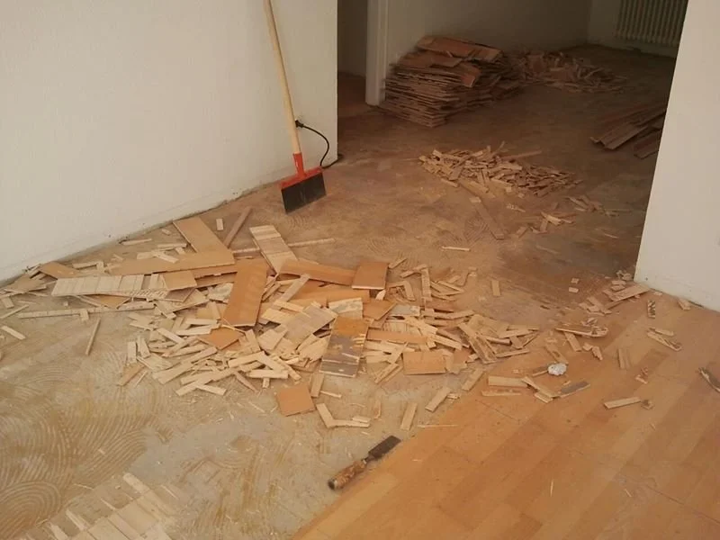 Zerbrochene Holz- oder Laminatfußbodenreste und ein Handbesen in einem Raum, vermutlich bei Renovierungsarbeiten.
