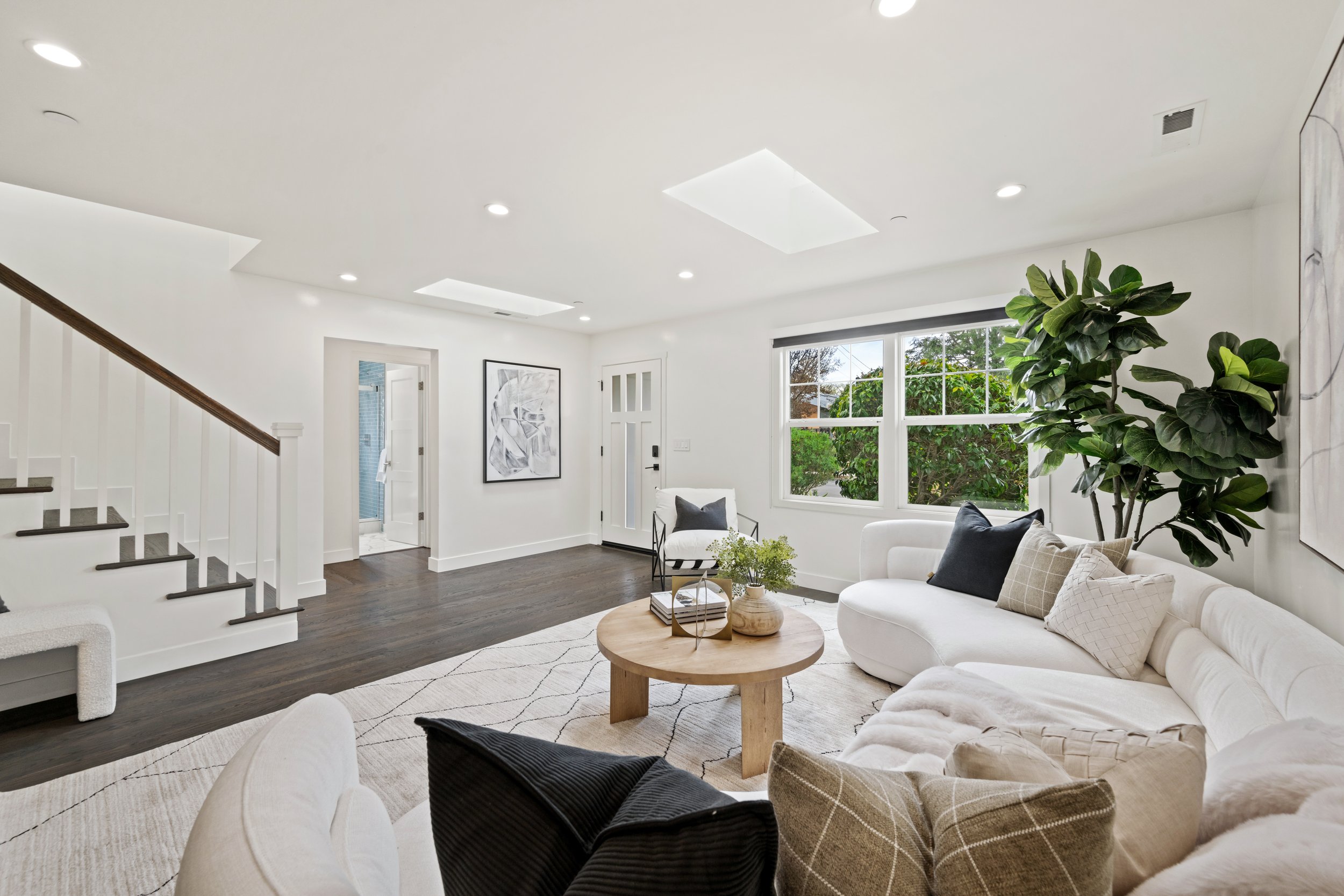 Living room with white walls, large windows, modern artwork, green plants, and beige sofas with pillows.
