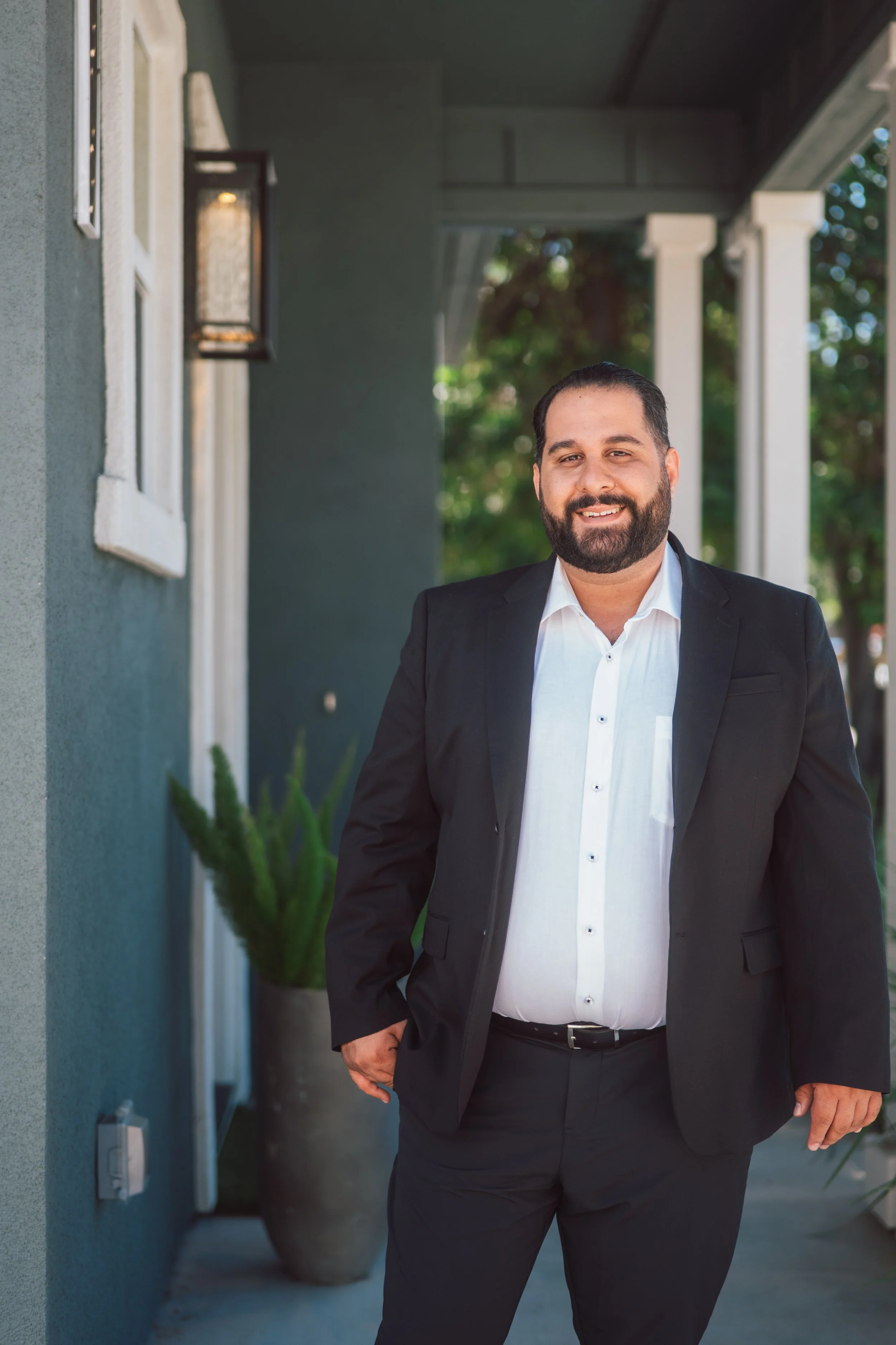 A man with a beard wearing a white shirt and black blazer smiling, standing outside in front of a grey building with a lantern and a potted plant.