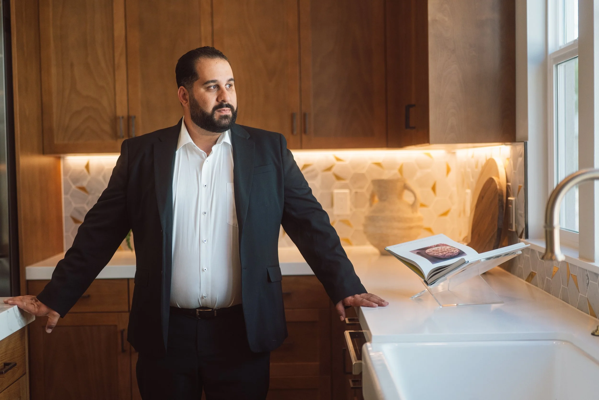 A man with a beard in a suit jacket and white shirt stands in a modern kitchen, leaning on a white countertop, with an open cookbook displaying a steak and vegetables.