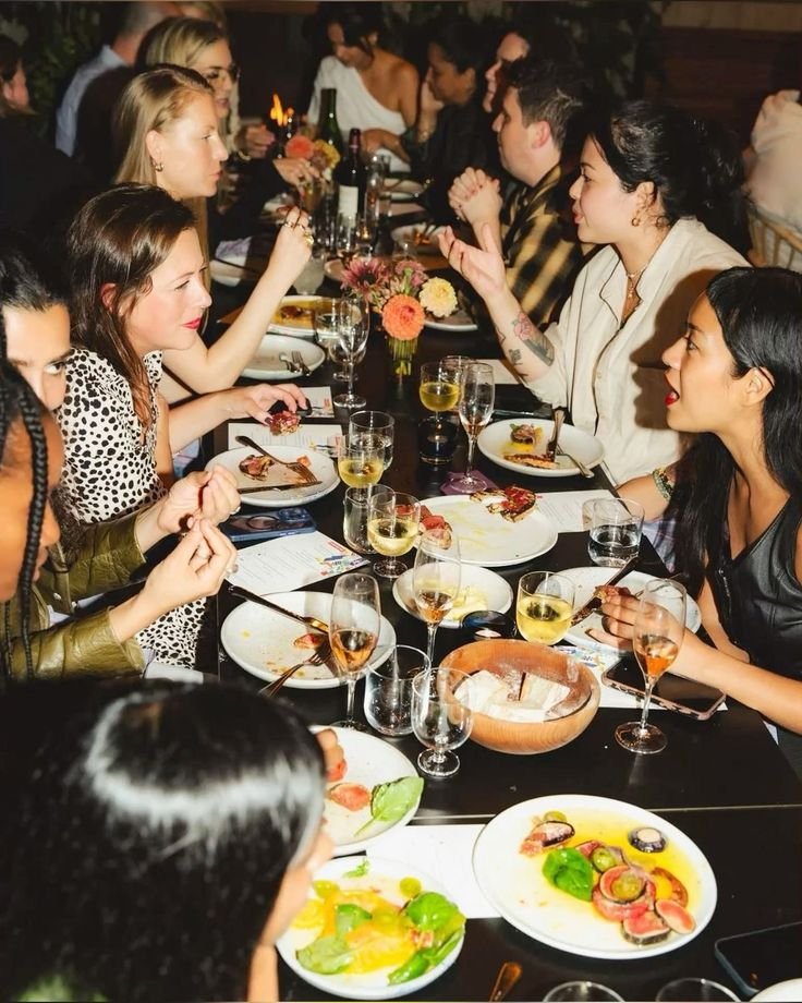 People dining at a long table, enjoying a meal with plates of food and glasses of wine in a dimly lit restaurant.