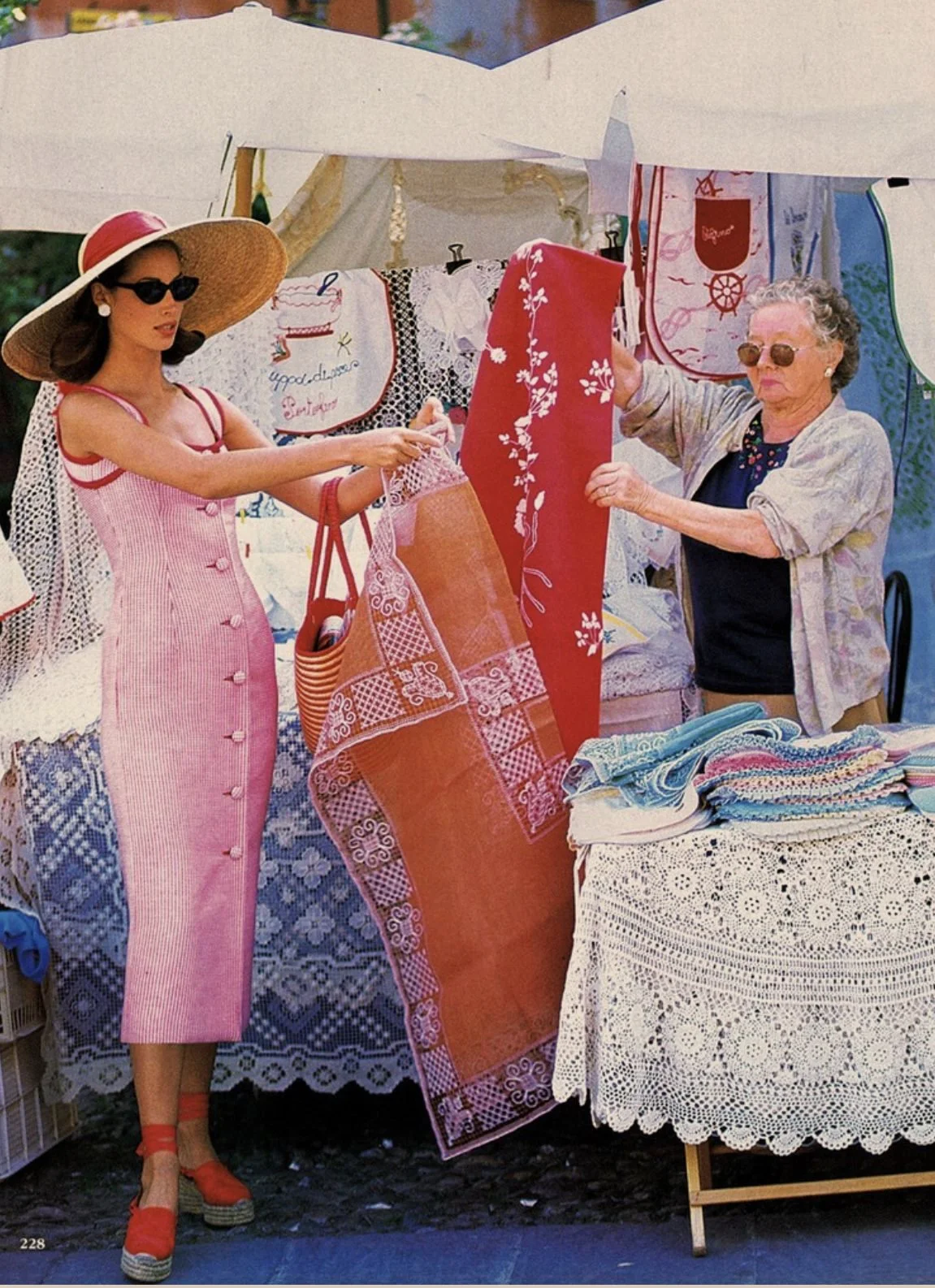 A woman in a pink dress with buttons, large sun hat, and sunglasses shopping at a fabric stall run by an older woman with curly gray hair, sunglasses, and a light jacket, on an outdoor market day.
