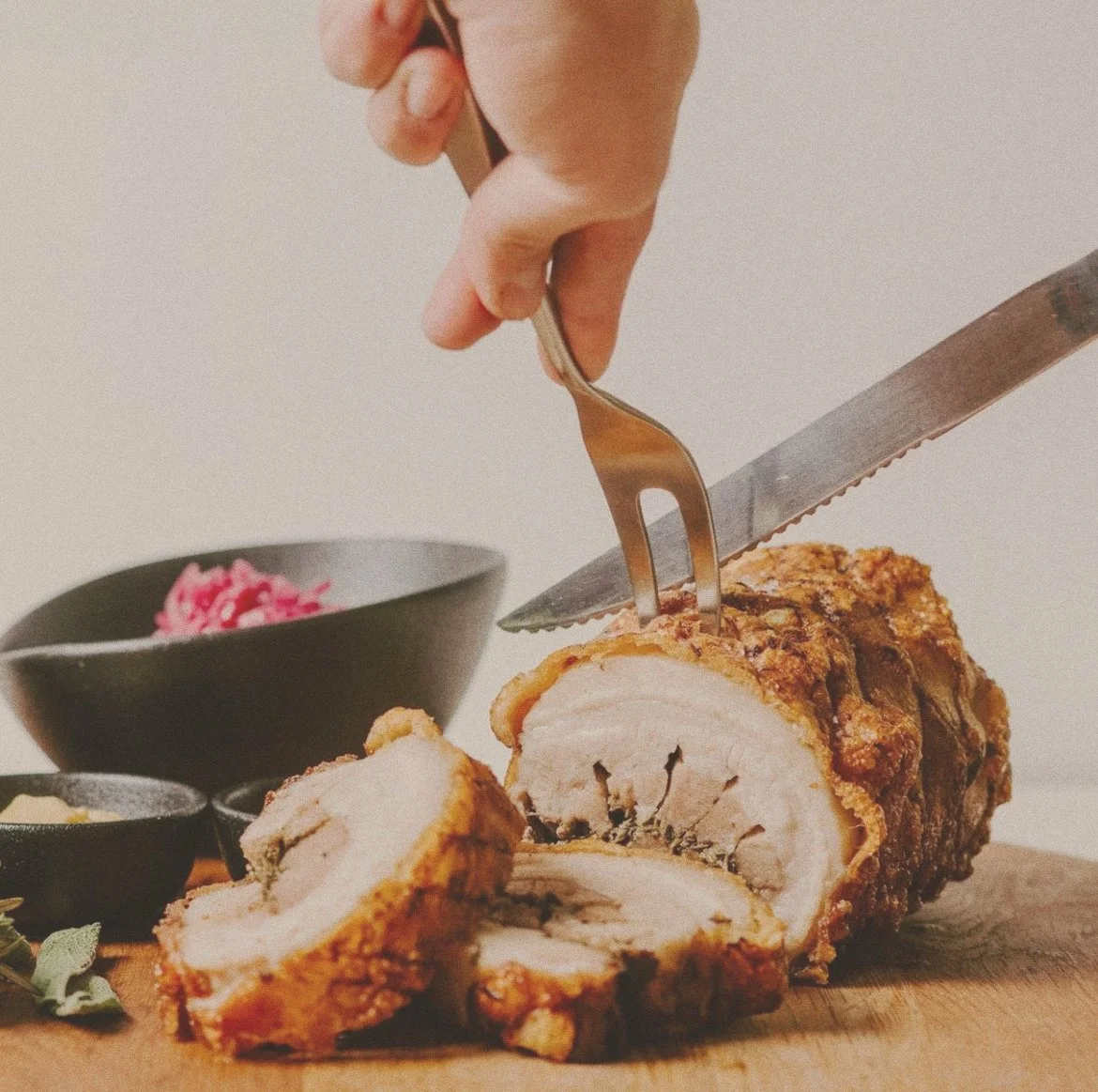Person slicing a cooked, breaded chicken roulade with a fork and knife on a wooden cutting board, with bowls of side dishes in the background.