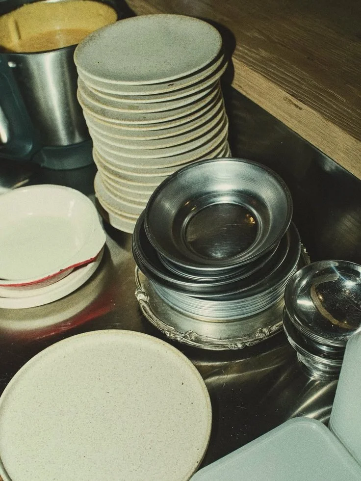 Stacked white ceramic plates, metal bowls, and small glass bowls on a dark countertop.