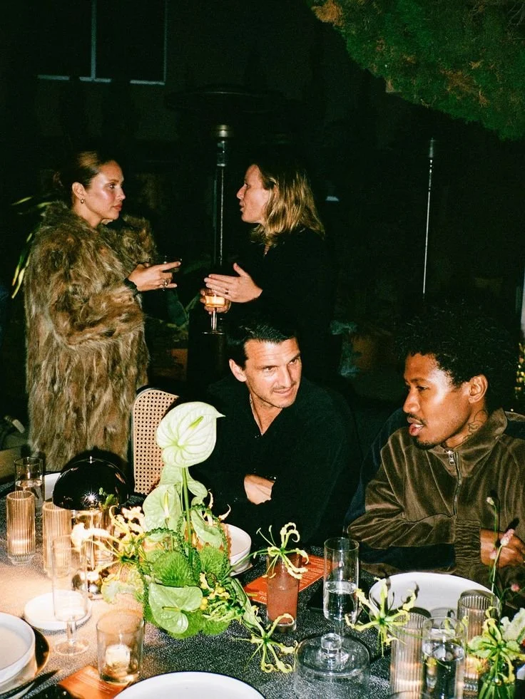 Four people are at a dinner table, two women are standing and two men are seated, with decorative flowers and tableware in the foreground.