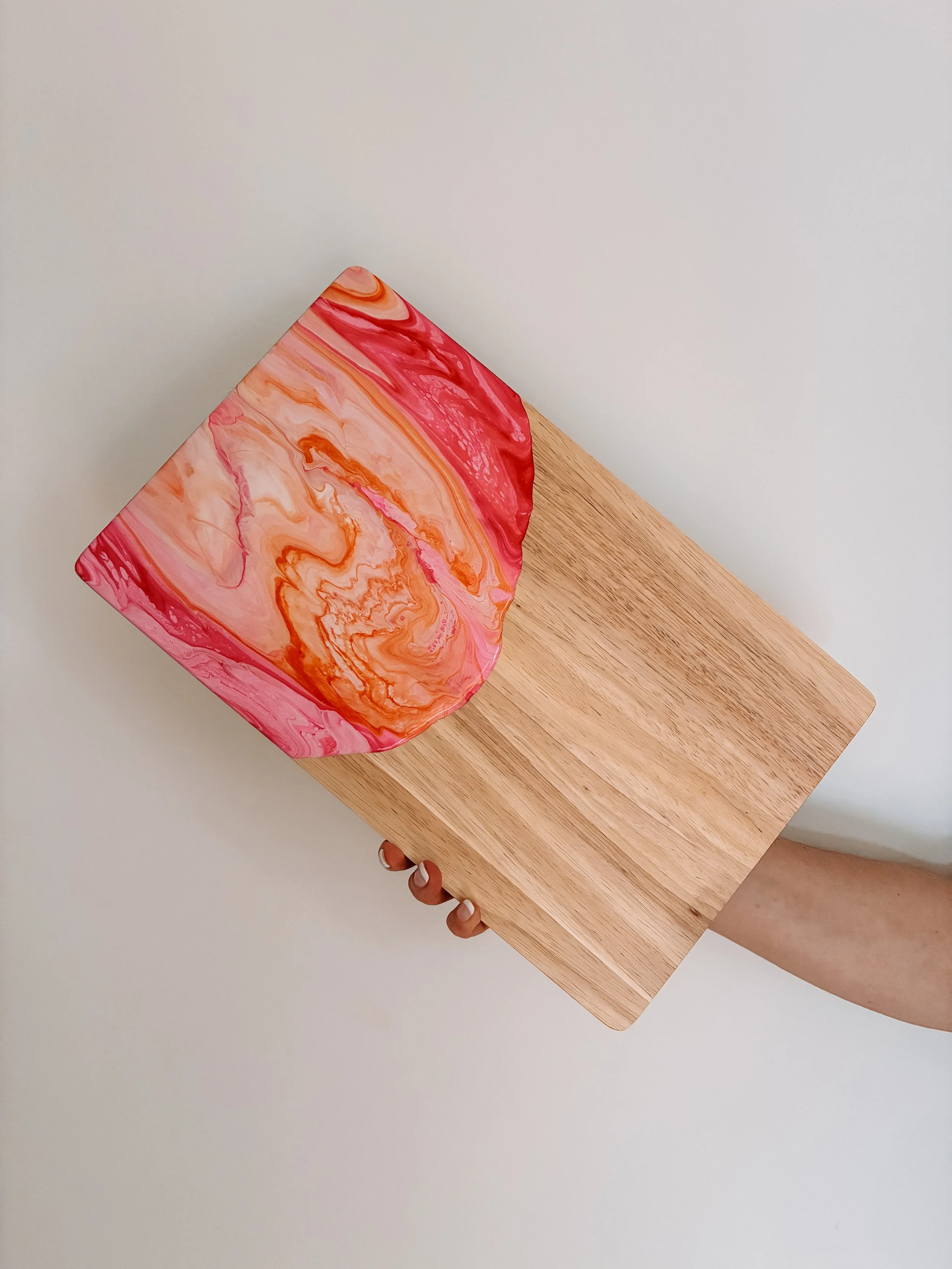 A person is holding a resin coated wooden cheeseboard with pink, orange, and white swirls, while standing against a white wall.