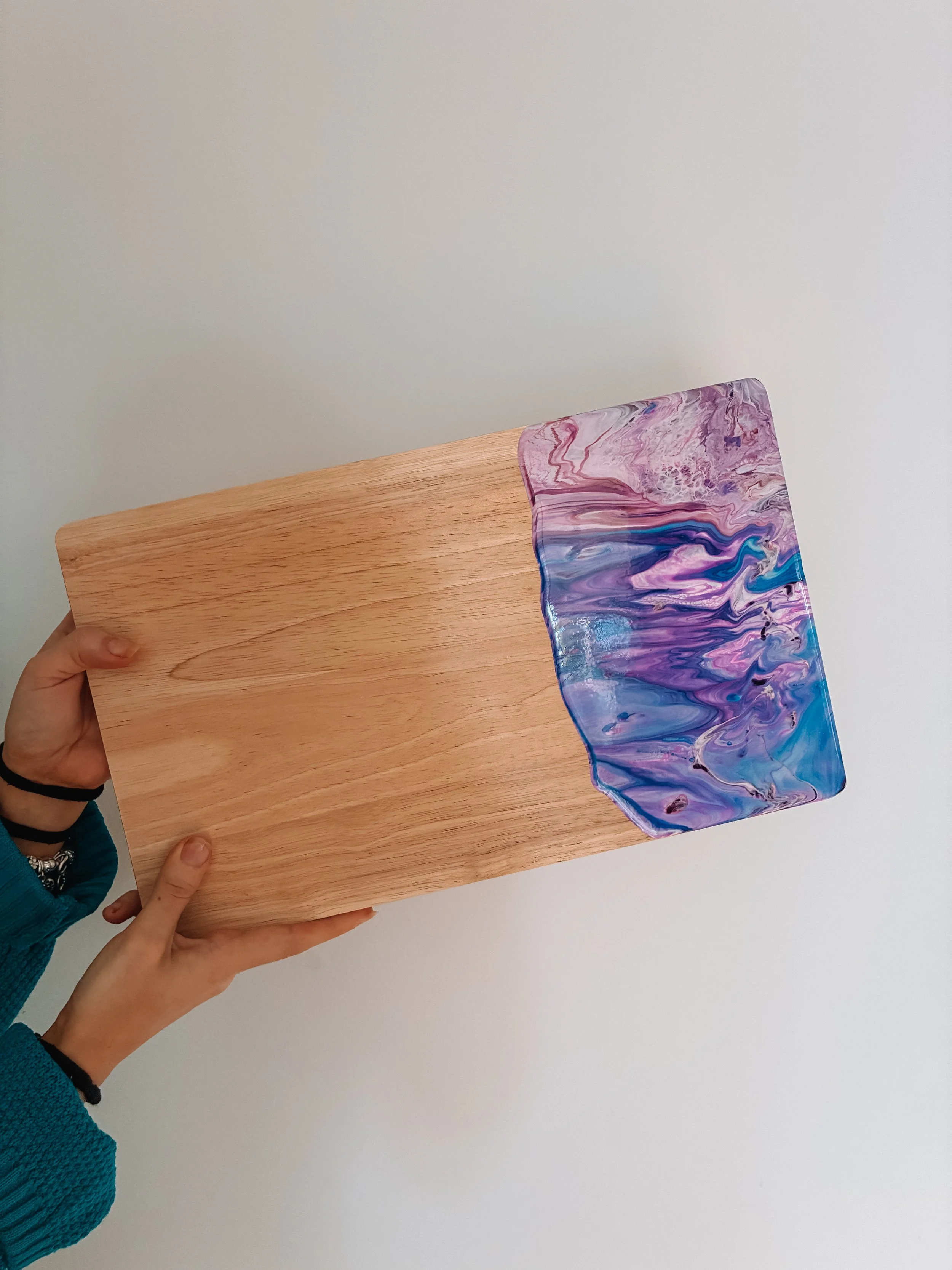Two hands holding a resin coated wooden cheeseboard with a purple and blue marbled pattern, against a plain white background.