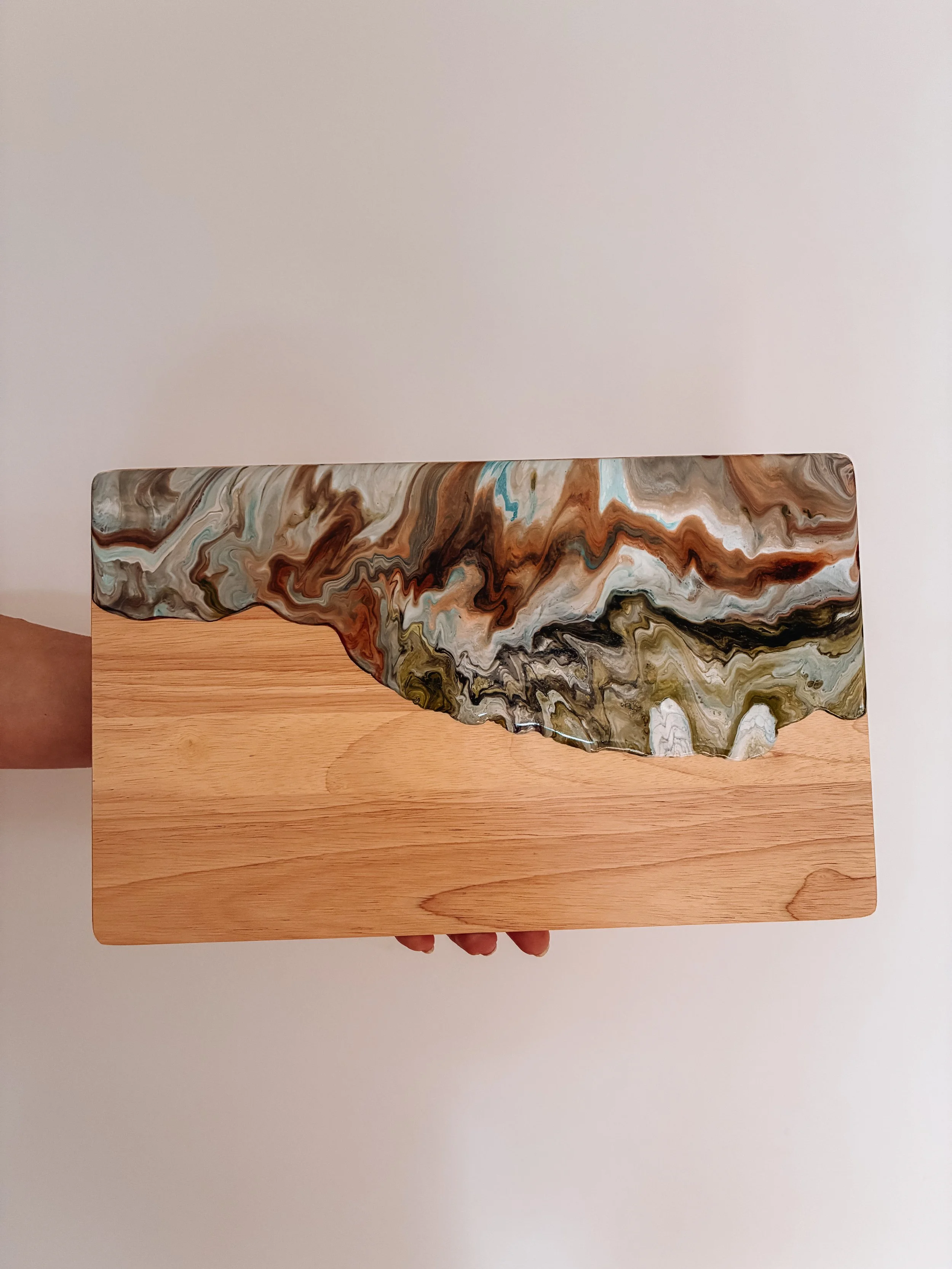 A person holding a resin coated wooden cheeseboard with swirling marble pattern in browns, blacks, and whites, over a white wall
