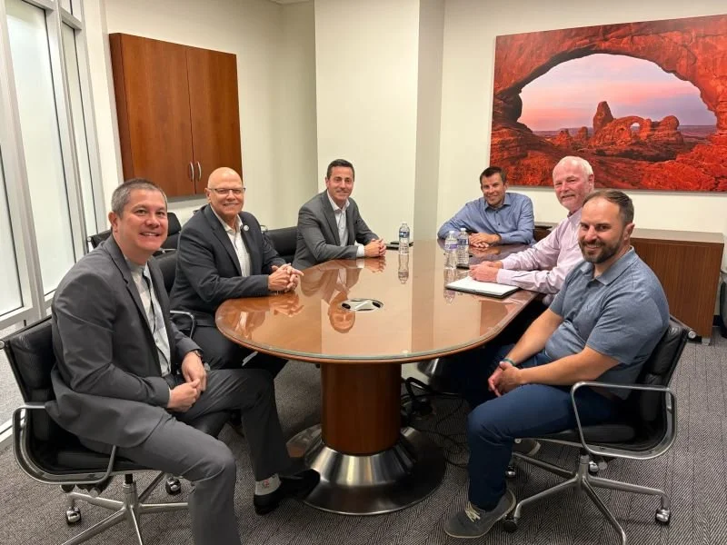 Six men in business attire sitting around a conference table in a meeting room, with a large landscape artwork on the wall.