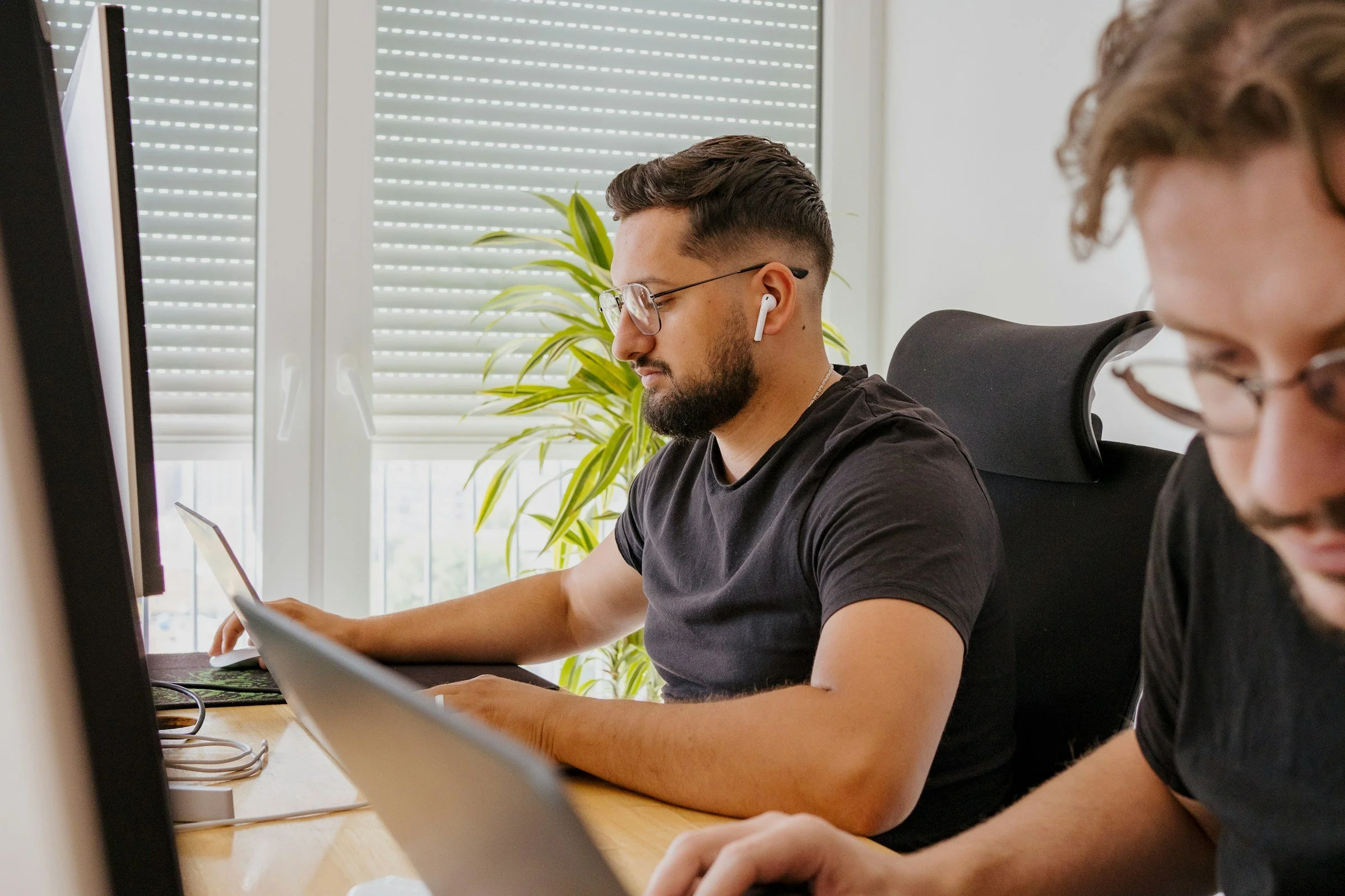 A man with glasses and wireless earbuds working at a desk with a laptop, with another person partially visible beside him, in a bright office with window blinds and a green plant in the background.