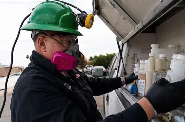 A worker wearing a green safety helmet, a pink respirator, and black gloves is handling bottles of chemicals or supplies from a service truck.