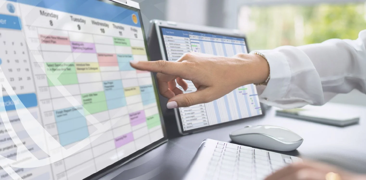 A person in a white shirt works at a desk with multiple screens displaying digital calendars and schedules, using their finger to interact with one of the screens.