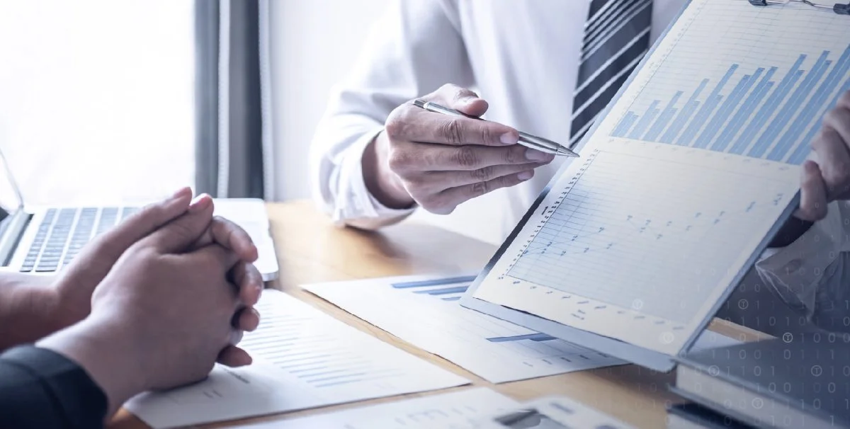 Two people discussing financial charts and graphs at a business meeting, light streaming in through the window.