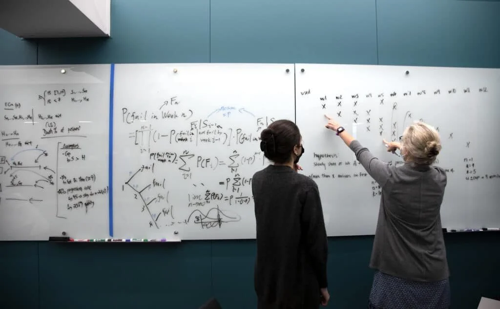 Two women are standing in front of a whiteboard filled with mathematical equations. One woman is pointing at the board, explaining something, while the other is listening.