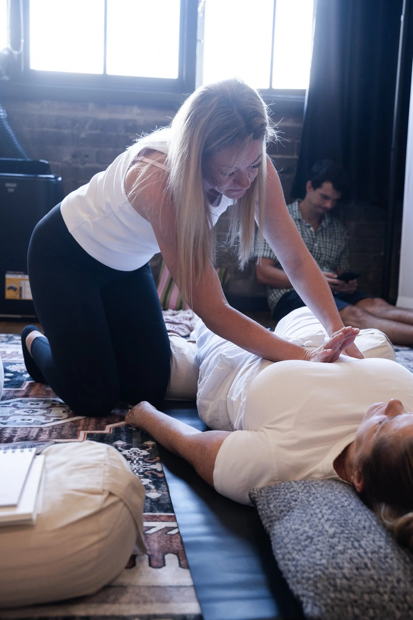 A woman facilitating transformational breathwork on a person lying on her back on the floor. The woman is kneeling beside her, applying intuitive touch with both hands.