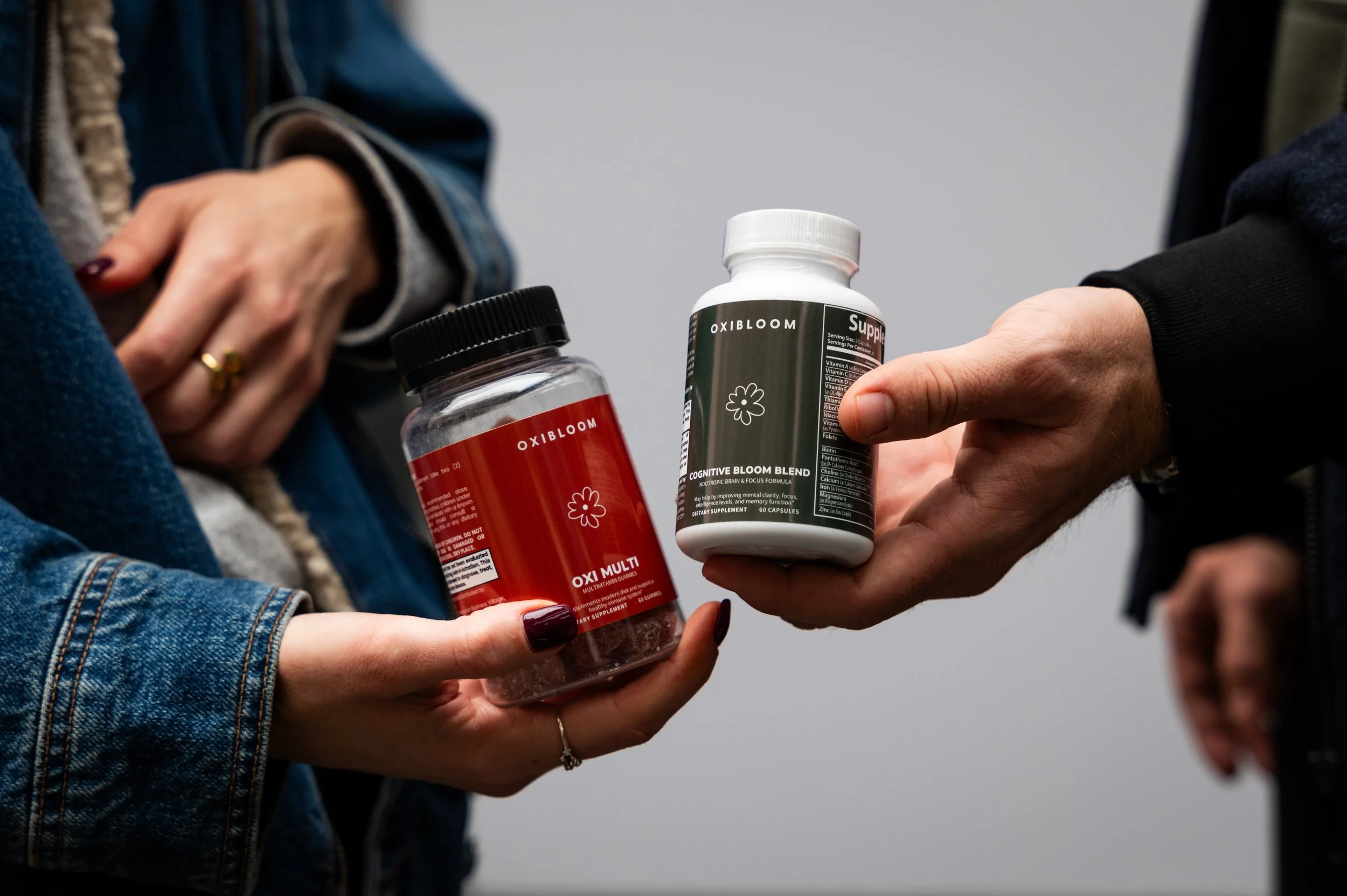 Two people exchanging bottles of dietary supplements, one red labeled 'OXIBLOOM' and the other green labeled 'OXIBLOOM'.