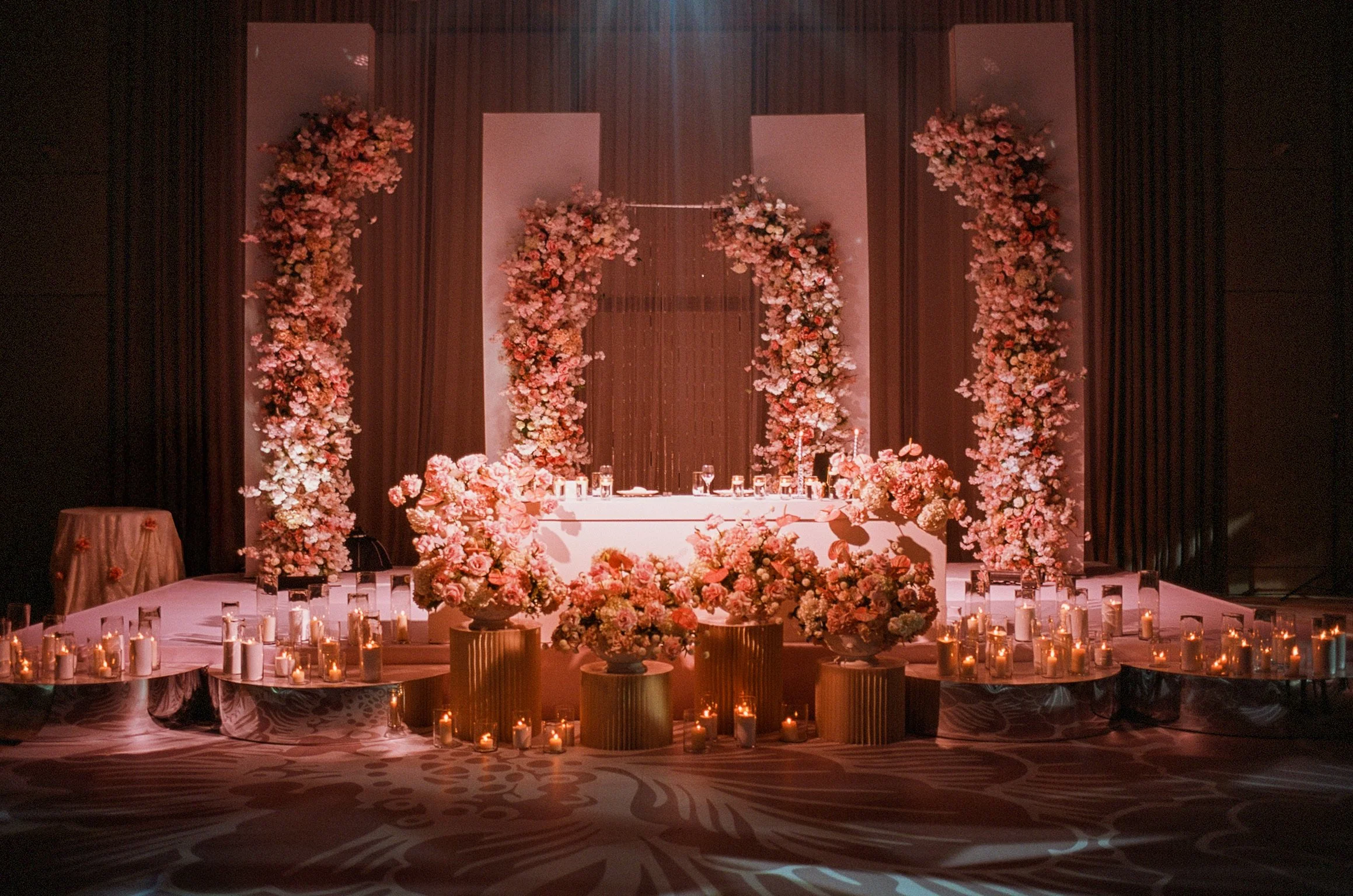 An elegant wedding setup with floral arrangements, candles, and a decorated table on a stage with dark curtains in the background.