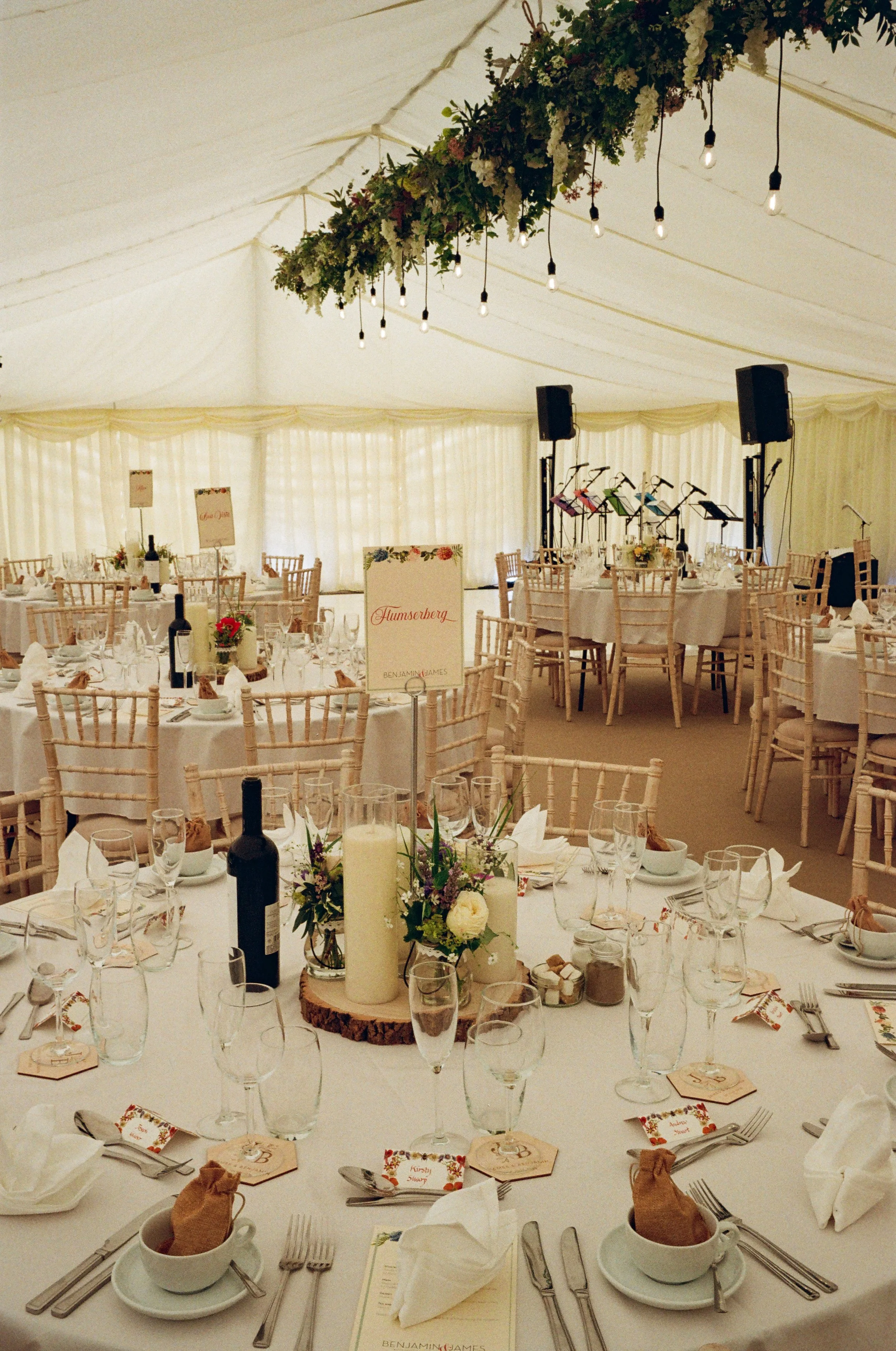 Round banquet tables set with white tablecloths, glassware, silverware, napkins, and place cards; a floral centerpiece with candles; a sign that says 'Humberley'; chairs arranged around the tables inside a large white tent decorated with string light