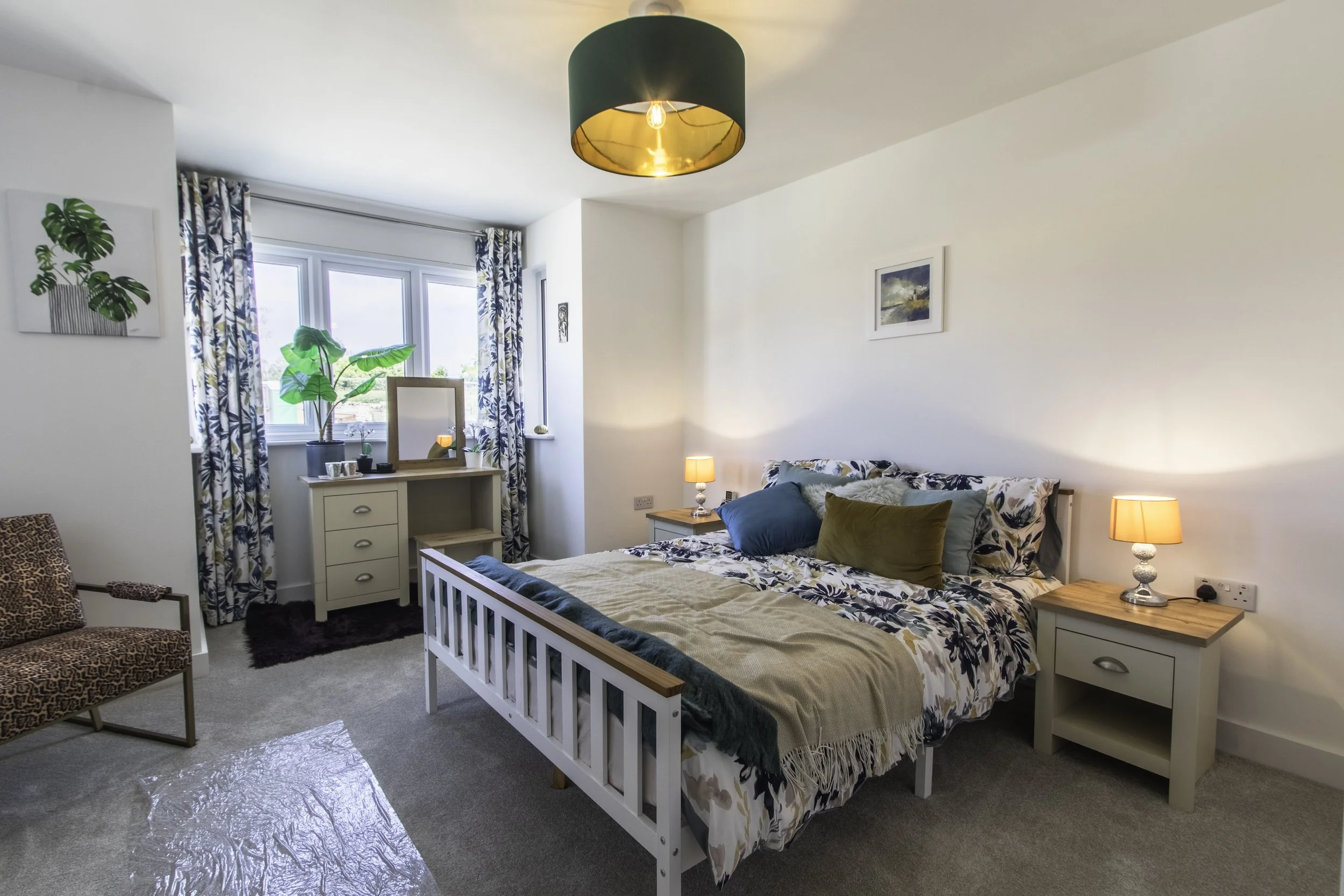 A bright bedroom with a white bed frame, patterned bedding, and a mix of colorful pillows. There are two nightstands with lamps, a small dresser with plants and decor, a window with floral curtains, and wall art. The ceiling has a black and gold pend