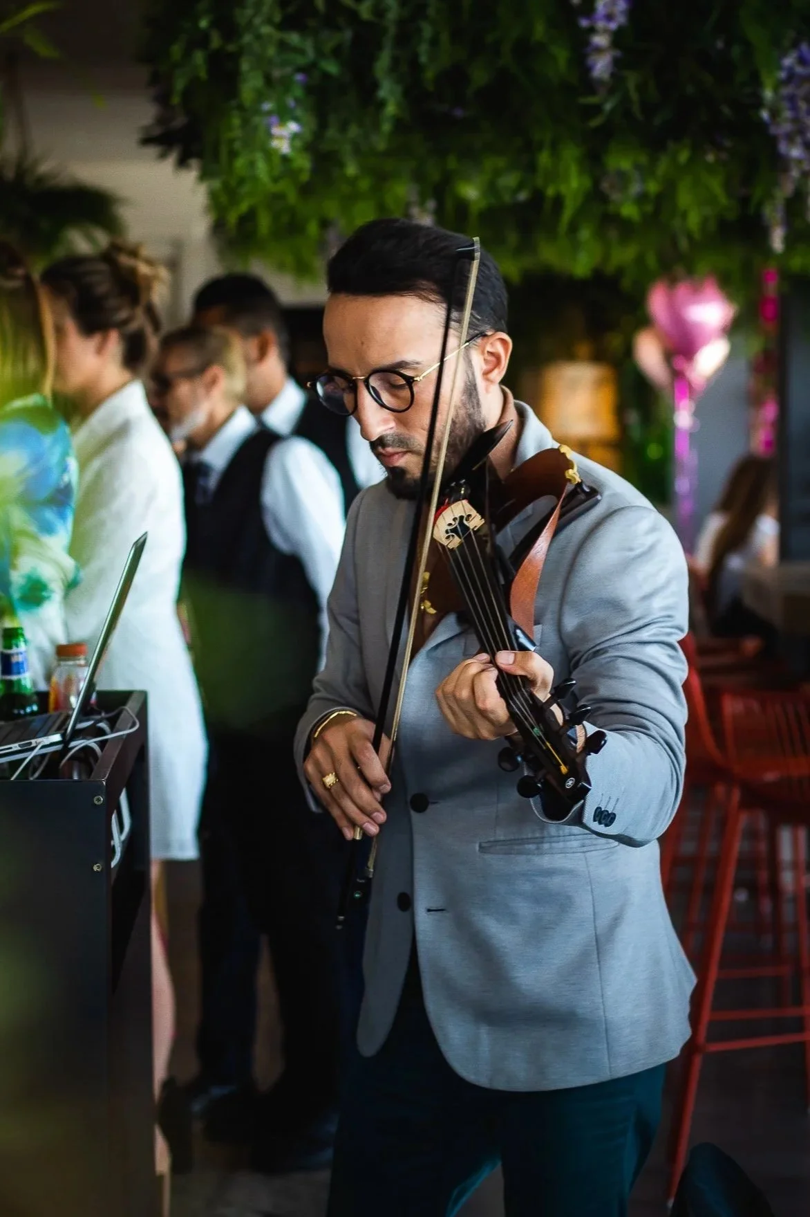 A man in a gray blazer playing a violin at an indoor event with green hanging plants and other people in the background.