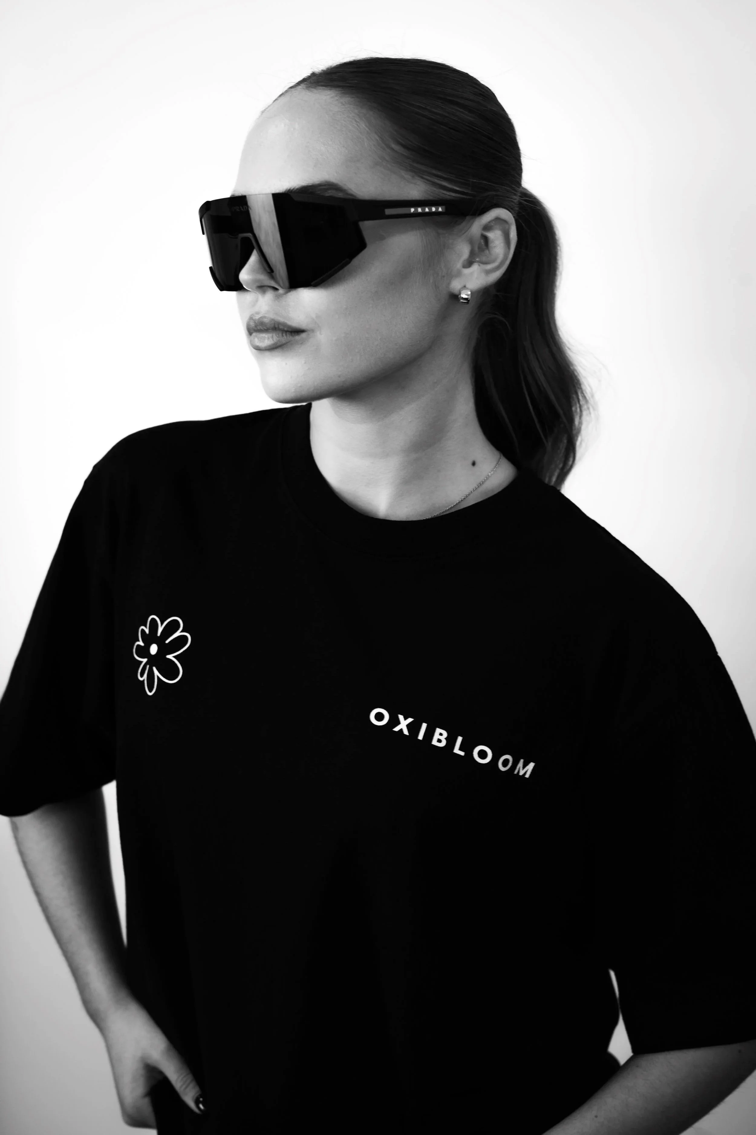 A woman wearing large black sunglasses with the Prada logo, a black T-shirt with a flower graphic, and the word 'OXIBLOOM' in white letters. Her hair is tied back, and she is standing against a plain white background.