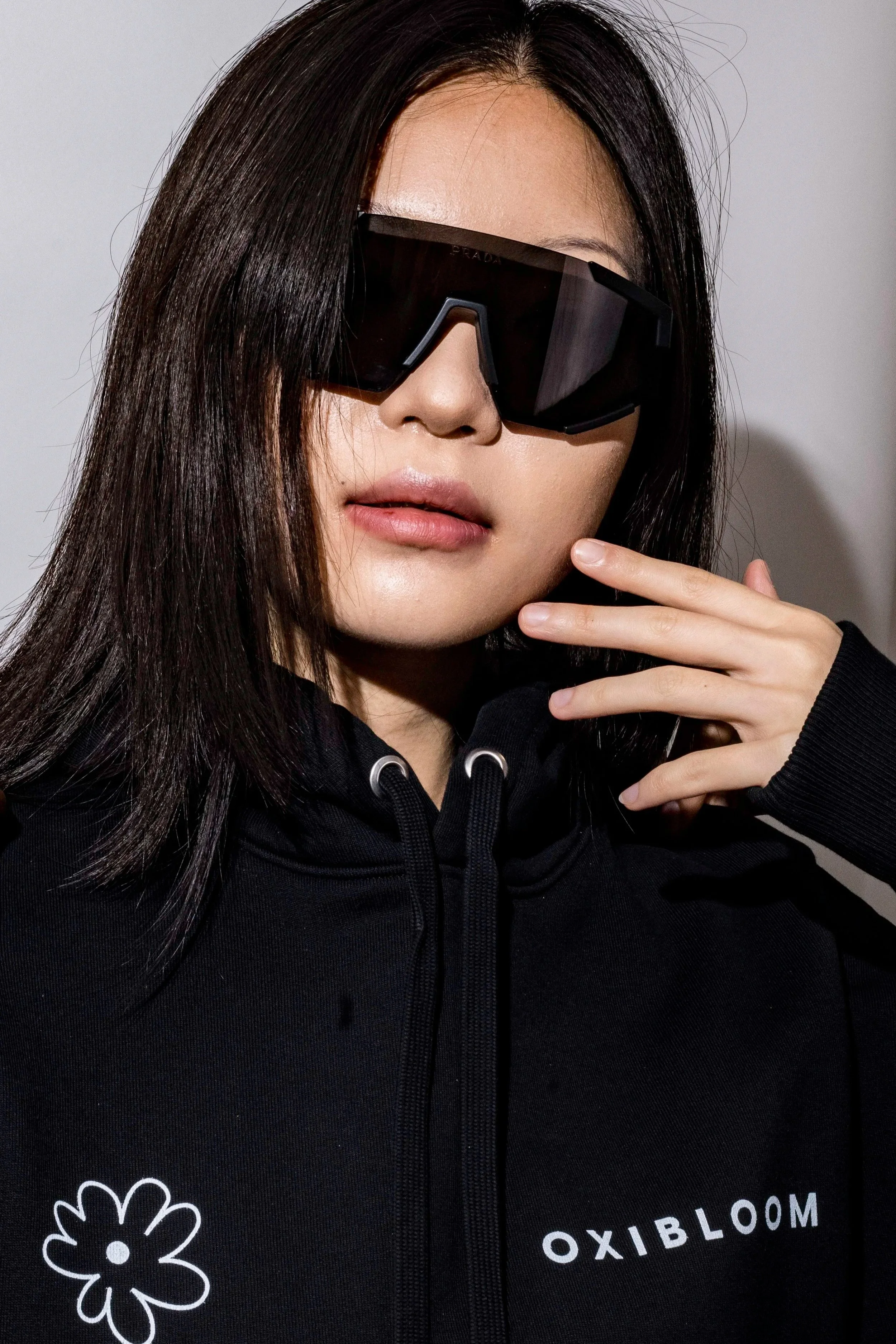 A woman with long dark hair wearing oversized black sunglasses and a black hoodie with a white flower logo and the text 'OXIBLOOM' on it.