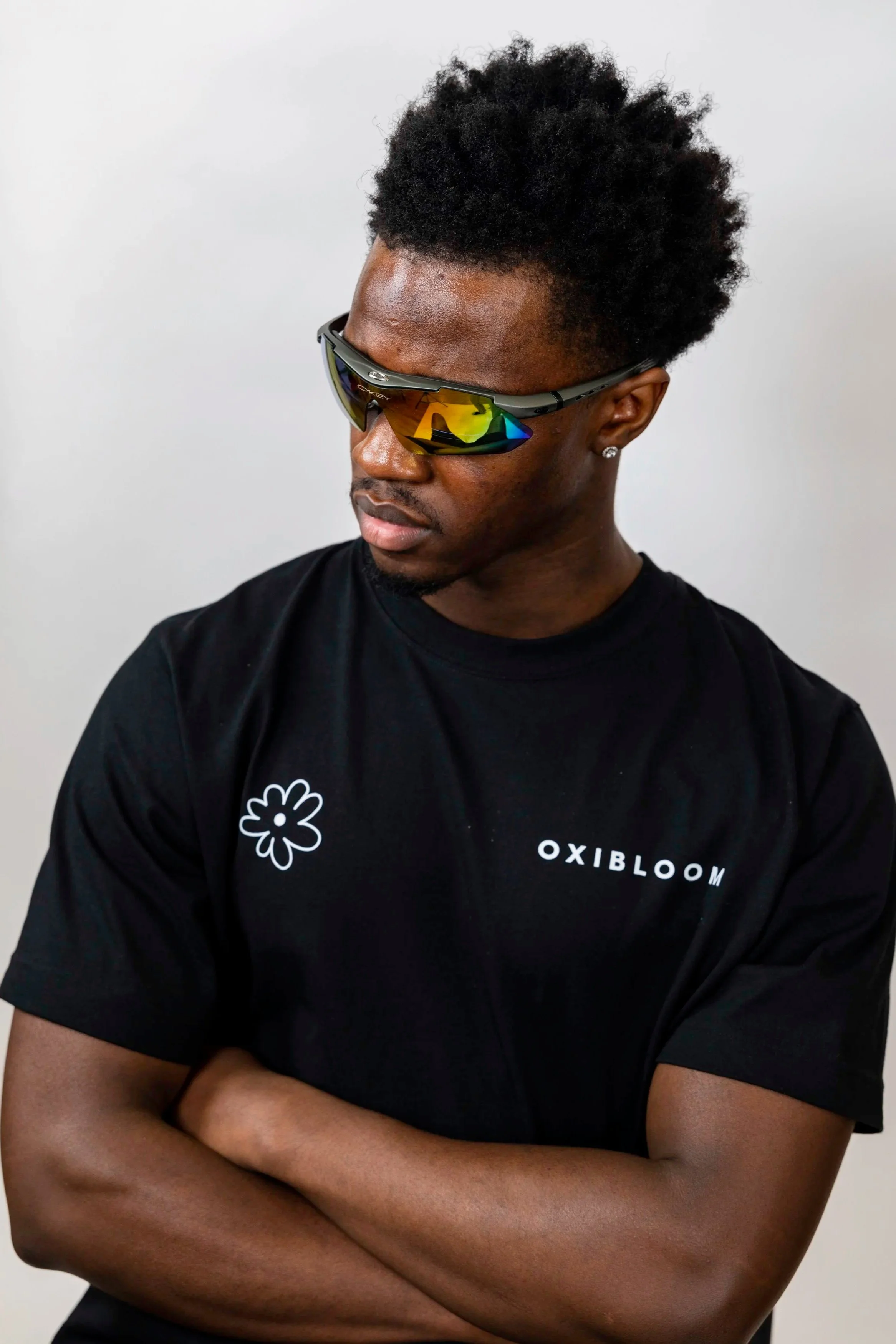 A young man with dark skin and curly black hair, wearing reflective sports sunglasses and a black T-shirt with a white flower logo and the word 'OXIBLOOM' on it. He has his arms crossed and is looking downward against a plain gray background.