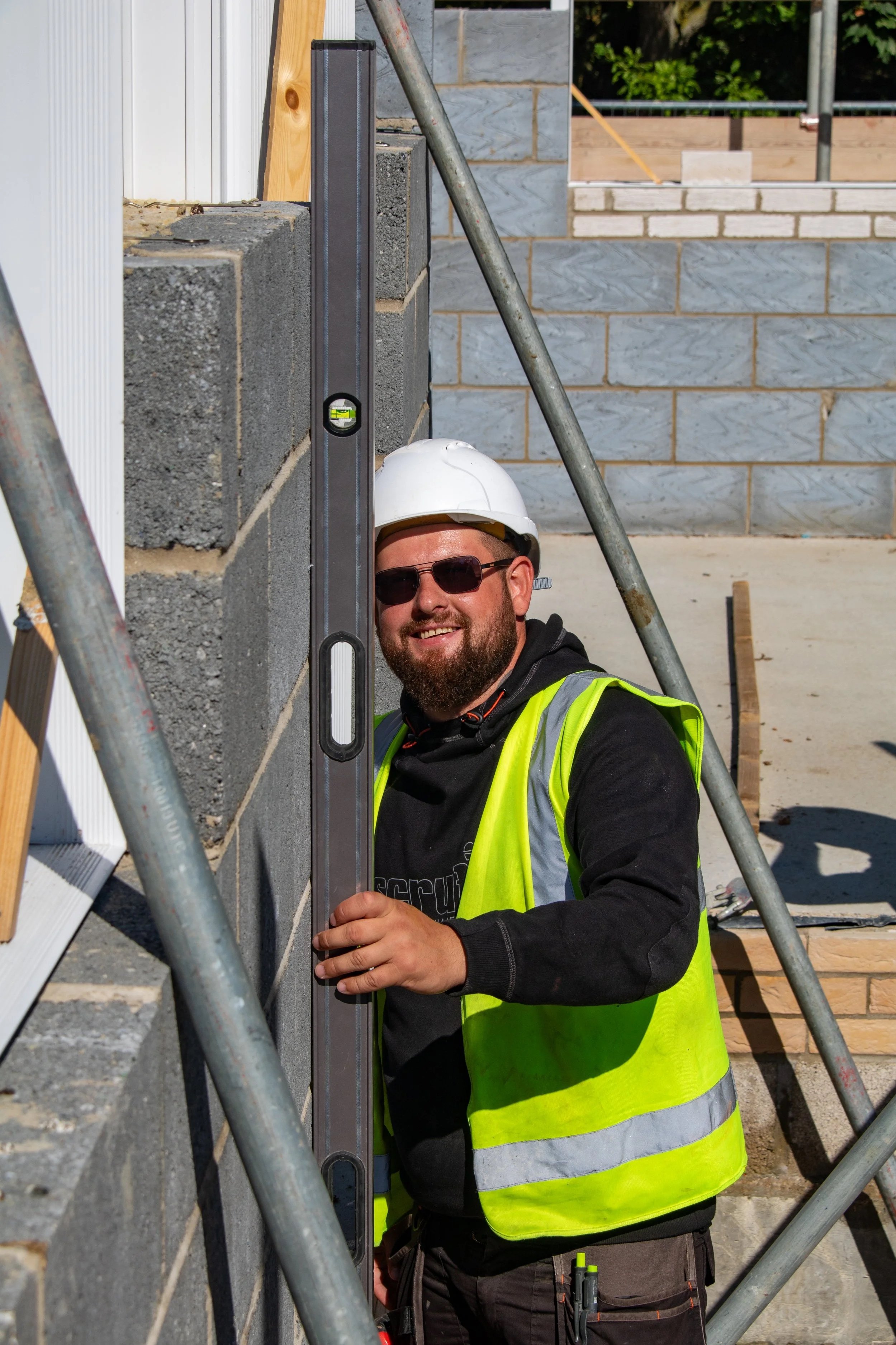 Construction worker with a safety helmet and sunglasses using a level on a brick wall during construction.