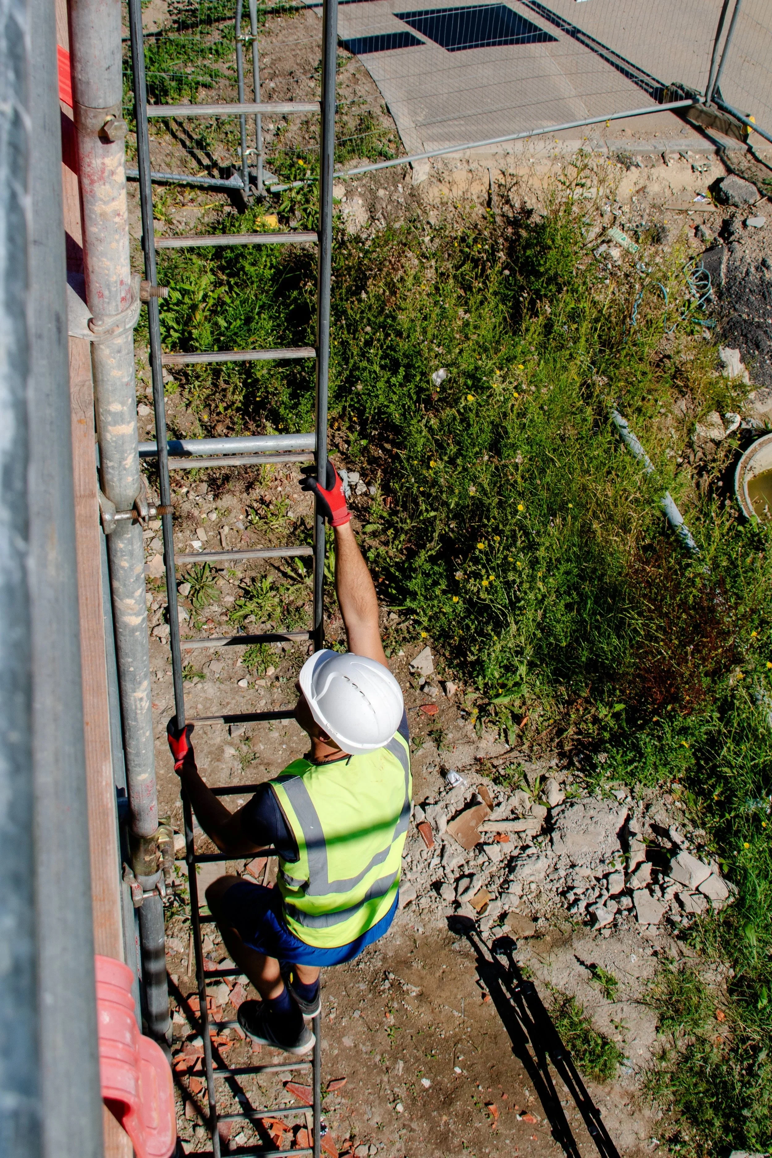 A construction worker wearing a white safety helmet, a yellow safety vest, and gloves is climbing a ladder at a construction site. The worker's right hand is gripping the ladder, and the ground around is rough and filled with rocks and debris. There 