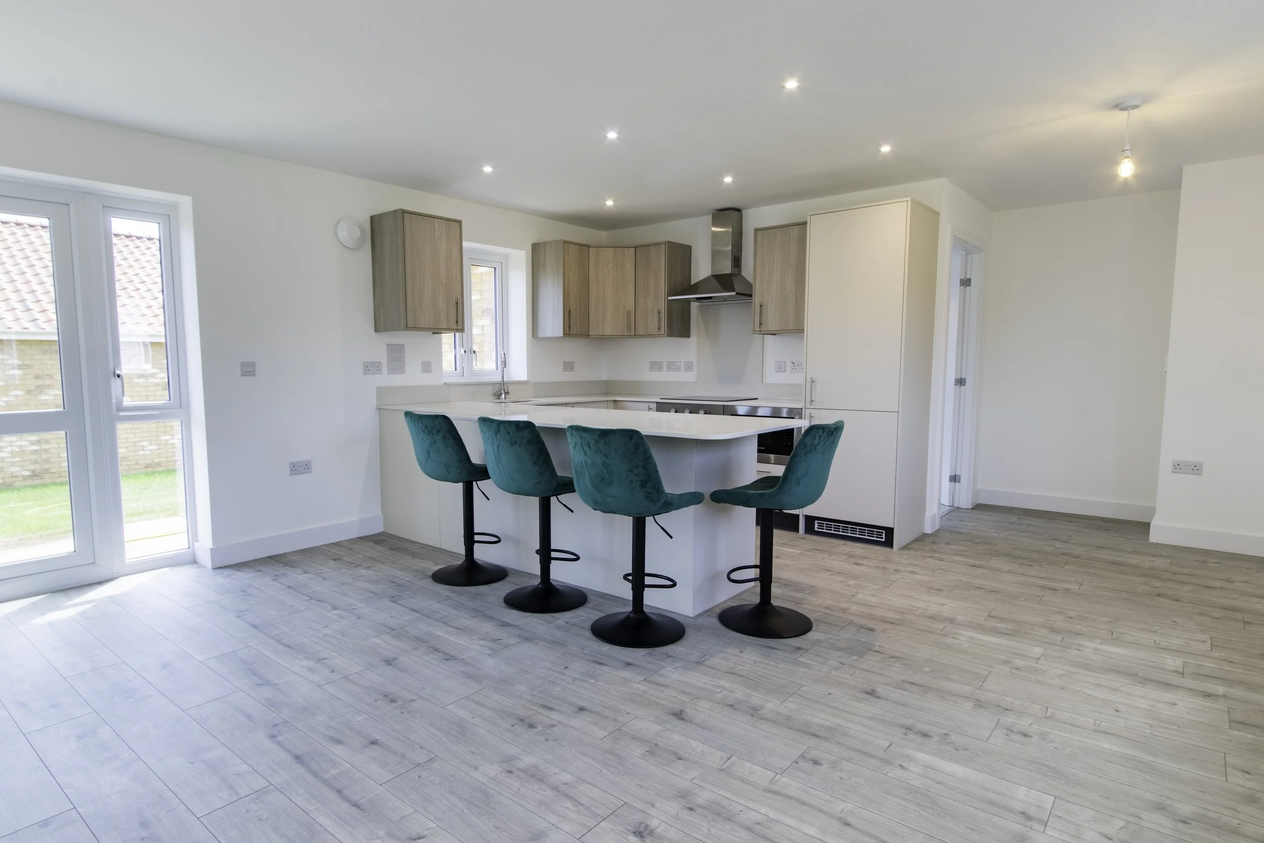 A modern kitchen with white walls, light wood cabinets, stainless steel appliances, a white countertop island with three teal bar stools, and a sliding glass door leading outside, under a ceiling with recessed lighting.