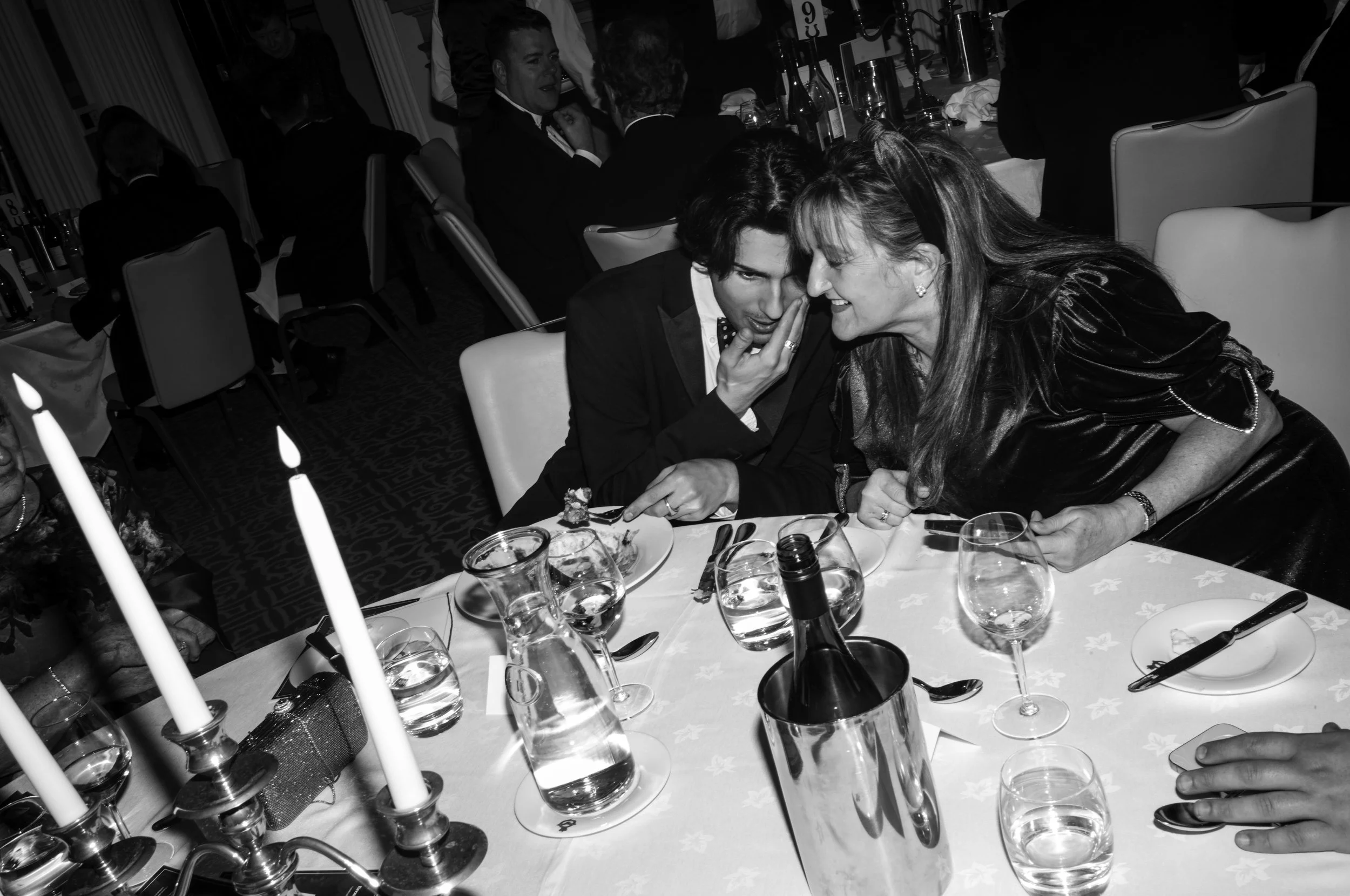 A group of people at a formal dinner gathering, with two women whispering and smiling at each other at the center of the image, surrounded by wine glasses, bottles, and table settings.