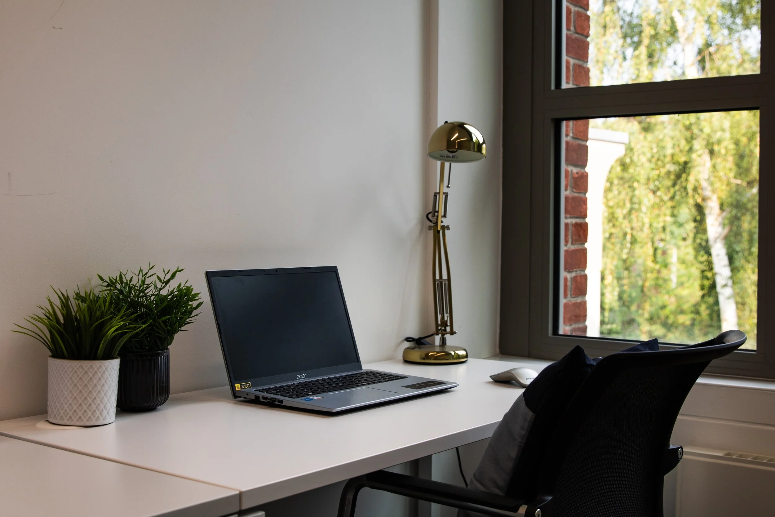 A modern office desk with a laptop, potted plants, a desk lamp, a computer mouse, and a chair near a large window showing trees outside.