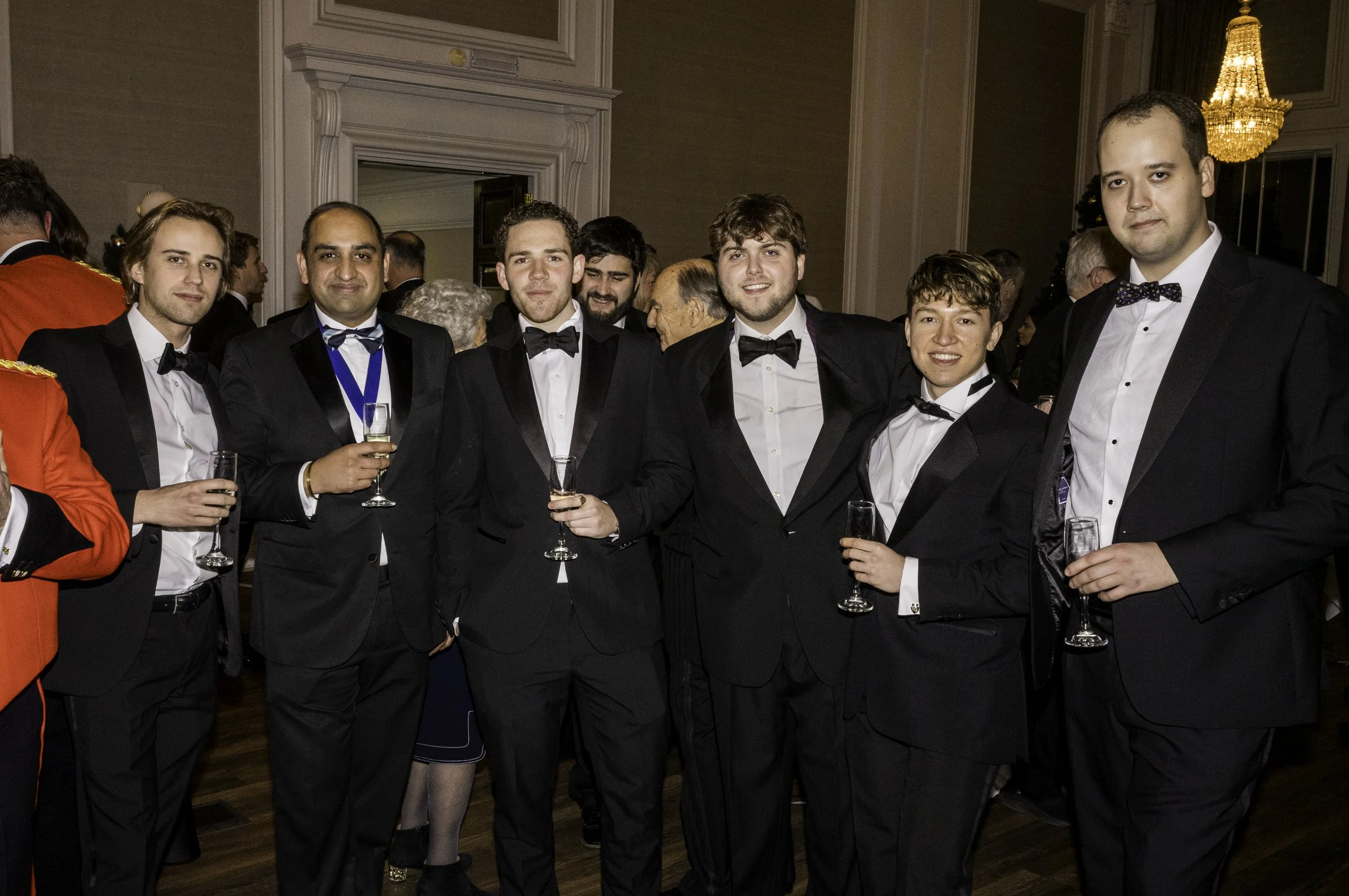 Group of six men in tuxedos posing at a formal event, holding champagne glasses, with a chandelier and other guests in the background.