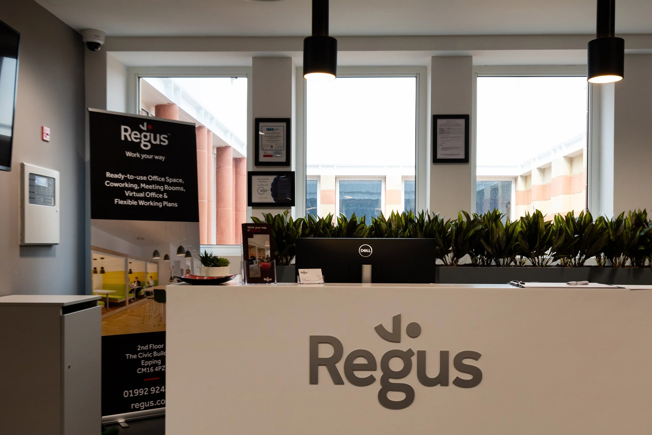 Office reception desk with a partial logo reading 'Regus' and promotional sign for office space and flexible working plans, large windows in the background letting in natural light, plants on the counter, and framed documents on the wall.
