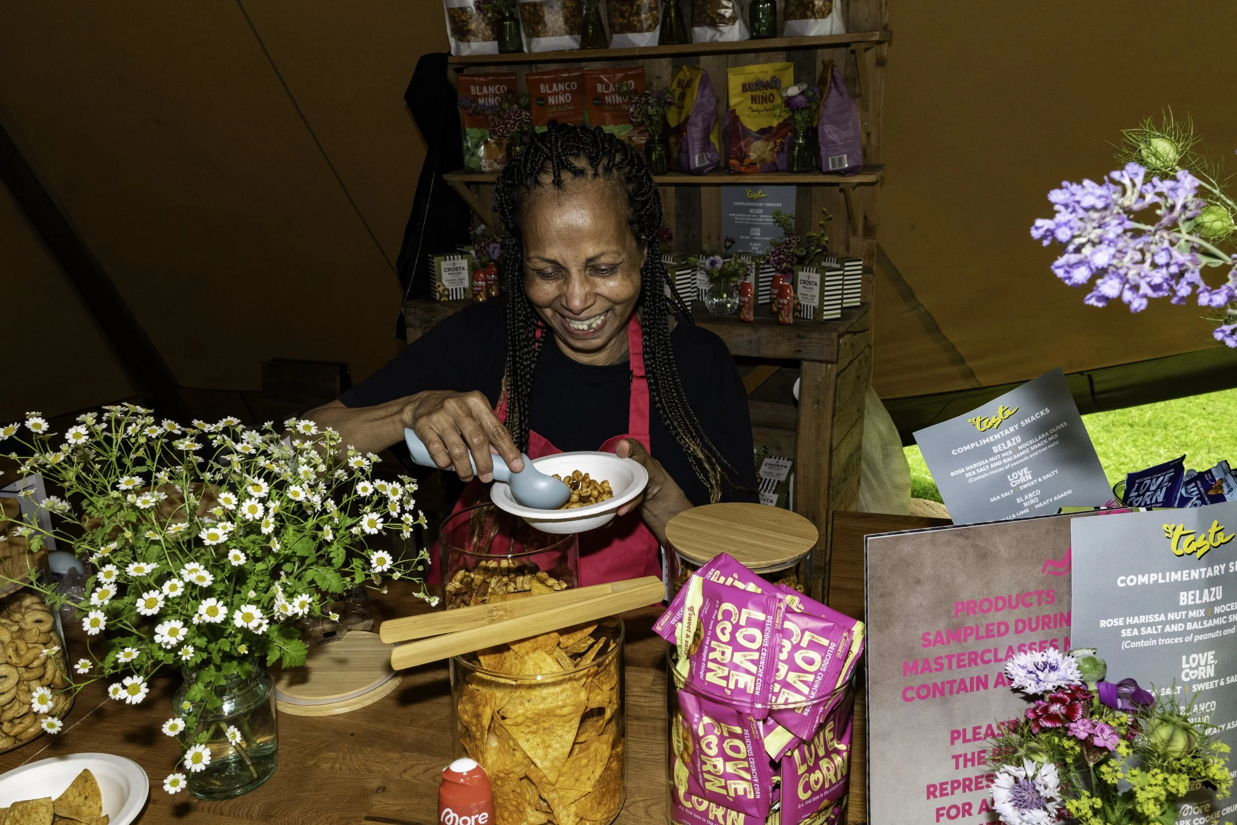 Woman smiling and serving food at a market stall with flowers, snack bags, and informational signs displayed.