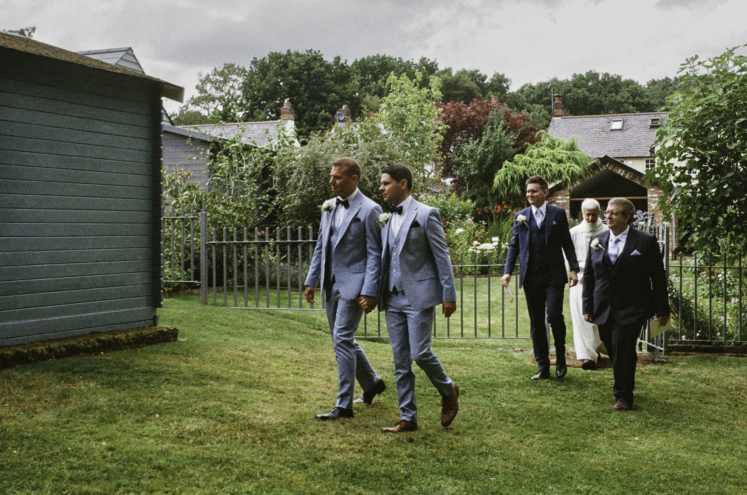 Group of men dressed in formal suits walking in a garden during a wedding or formal event.