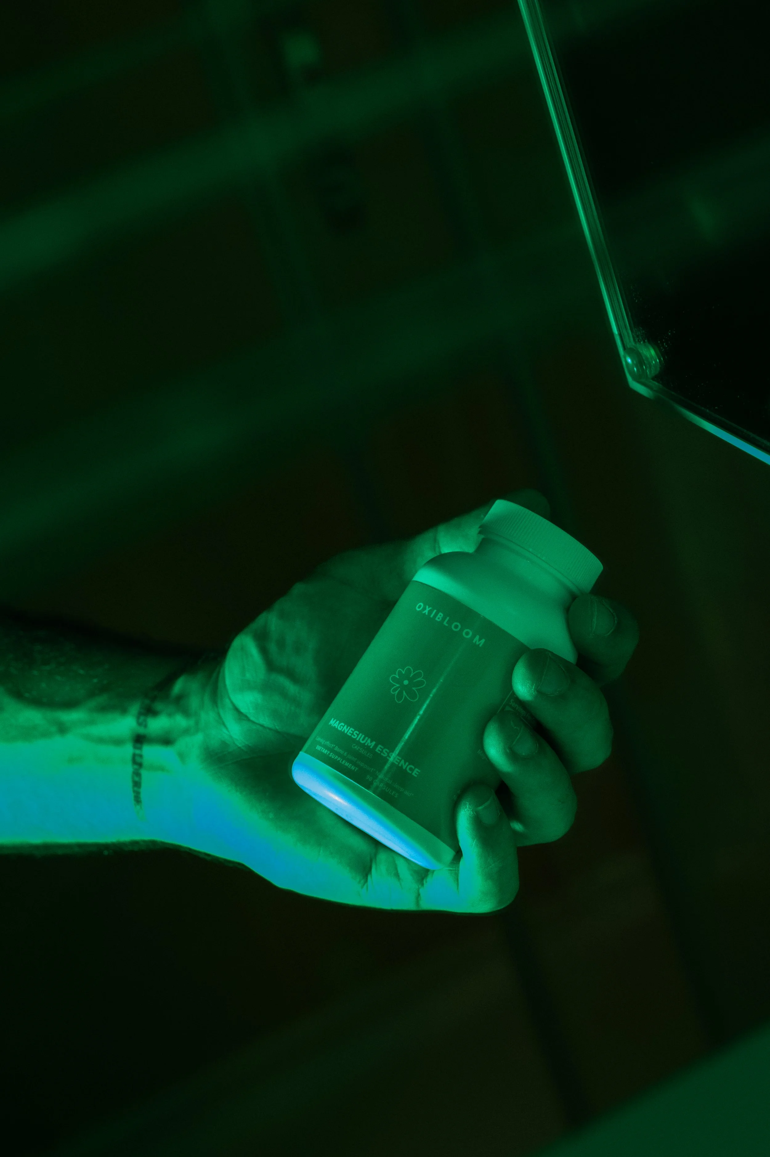A hand holding a bottle of magnesium supplement in front of a green light. The label on the bottle reads "Oxbloom Magnesium Essence."