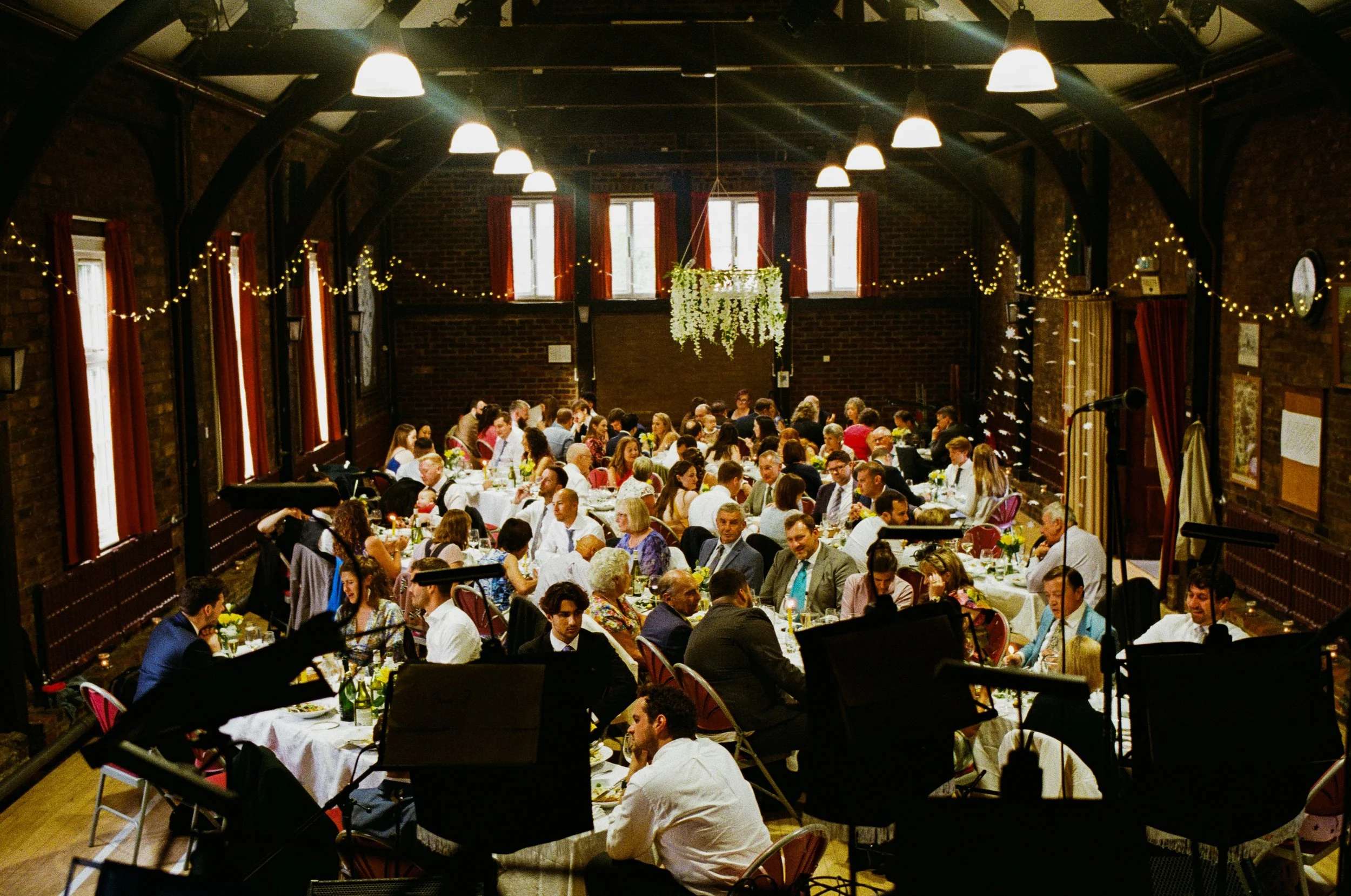 A large indoor gathering in a decorated hall with brick walls, string lights, and multiple long tables filled with people dining and socializing. The hall has high ceilings with exposed beams, and windows with red curtains.