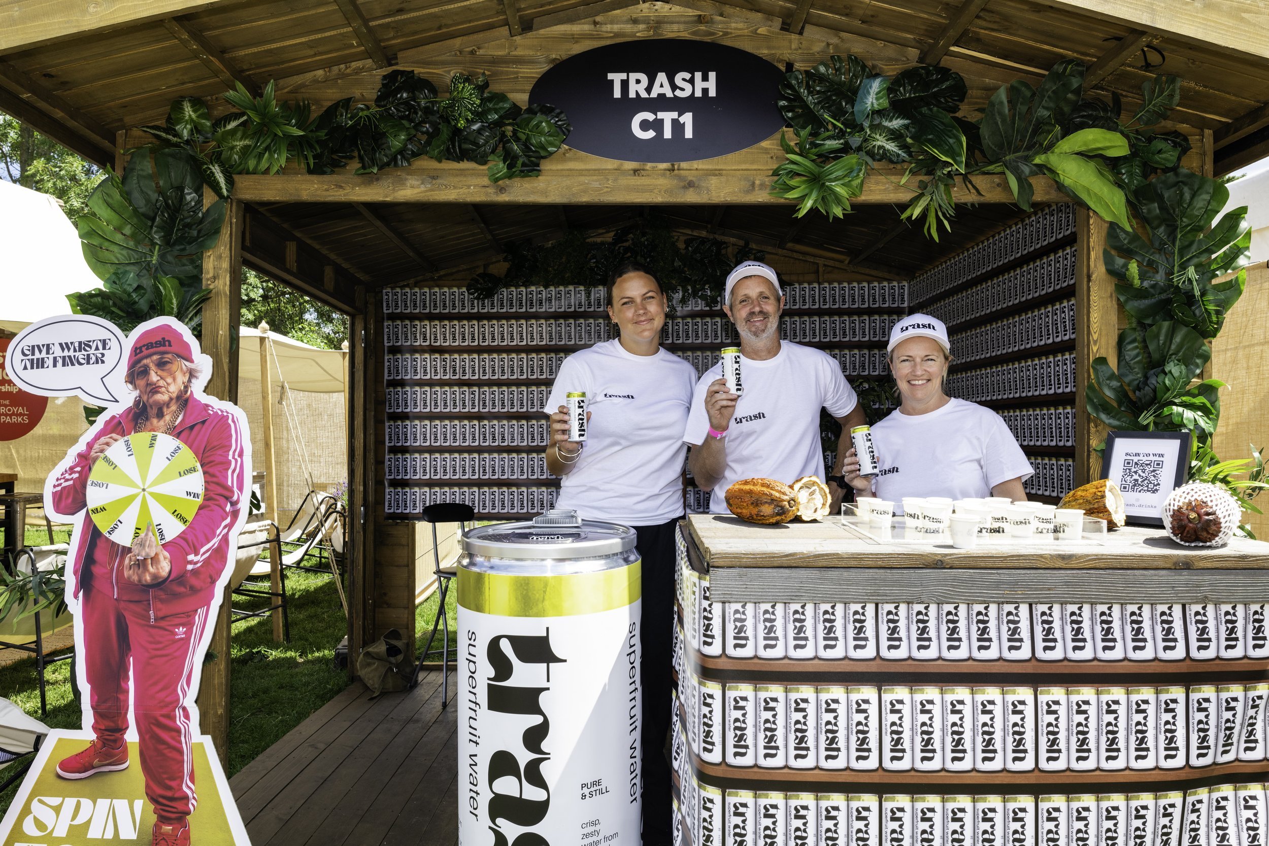 Group of three people standing behind a booth labeled 'Trash CT1' at an outdoor event, holding canned beverages and smiling. The booth is decorated with green foliage and has shelves filled with cans. To the left, there's a cardboard standee of an ol