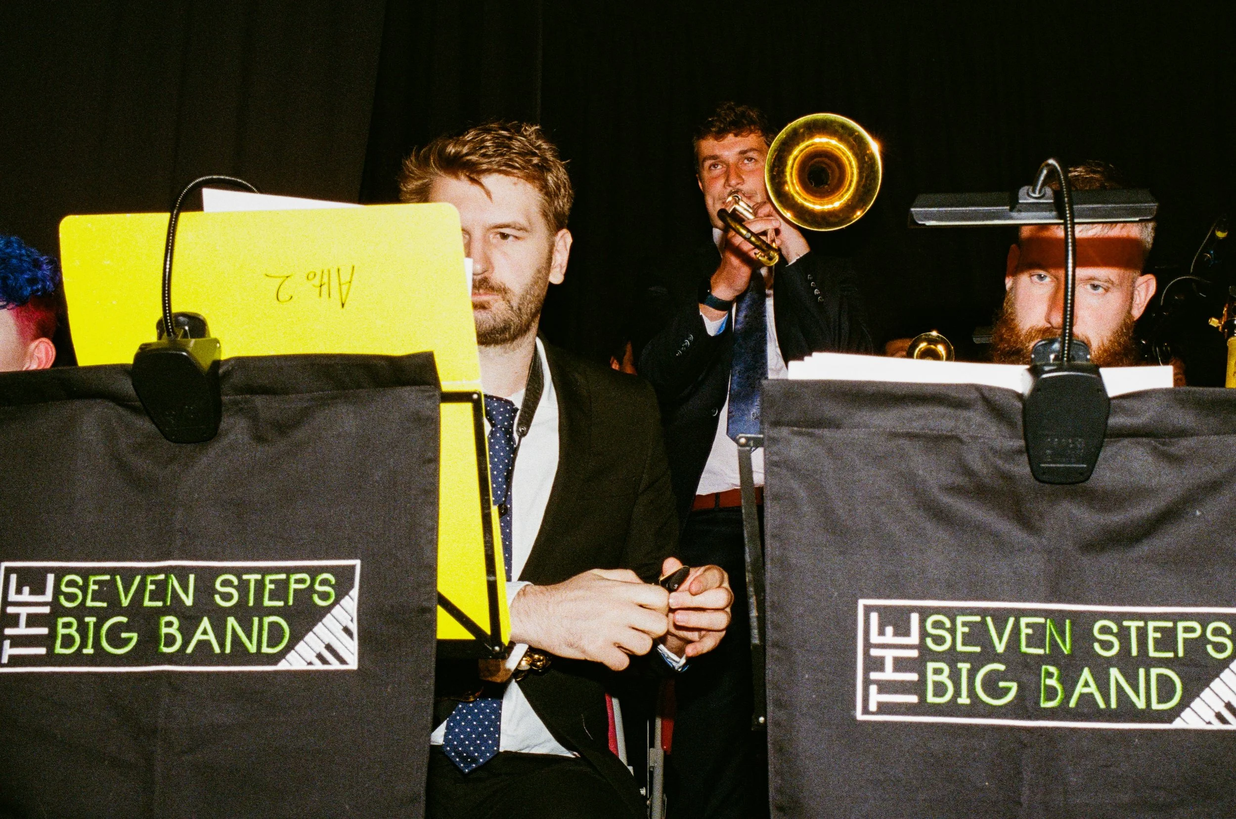 Musicians performing with music stands labeled 'The Seven Steps Big Band' on stage, with one man playing trumpet and others seated.