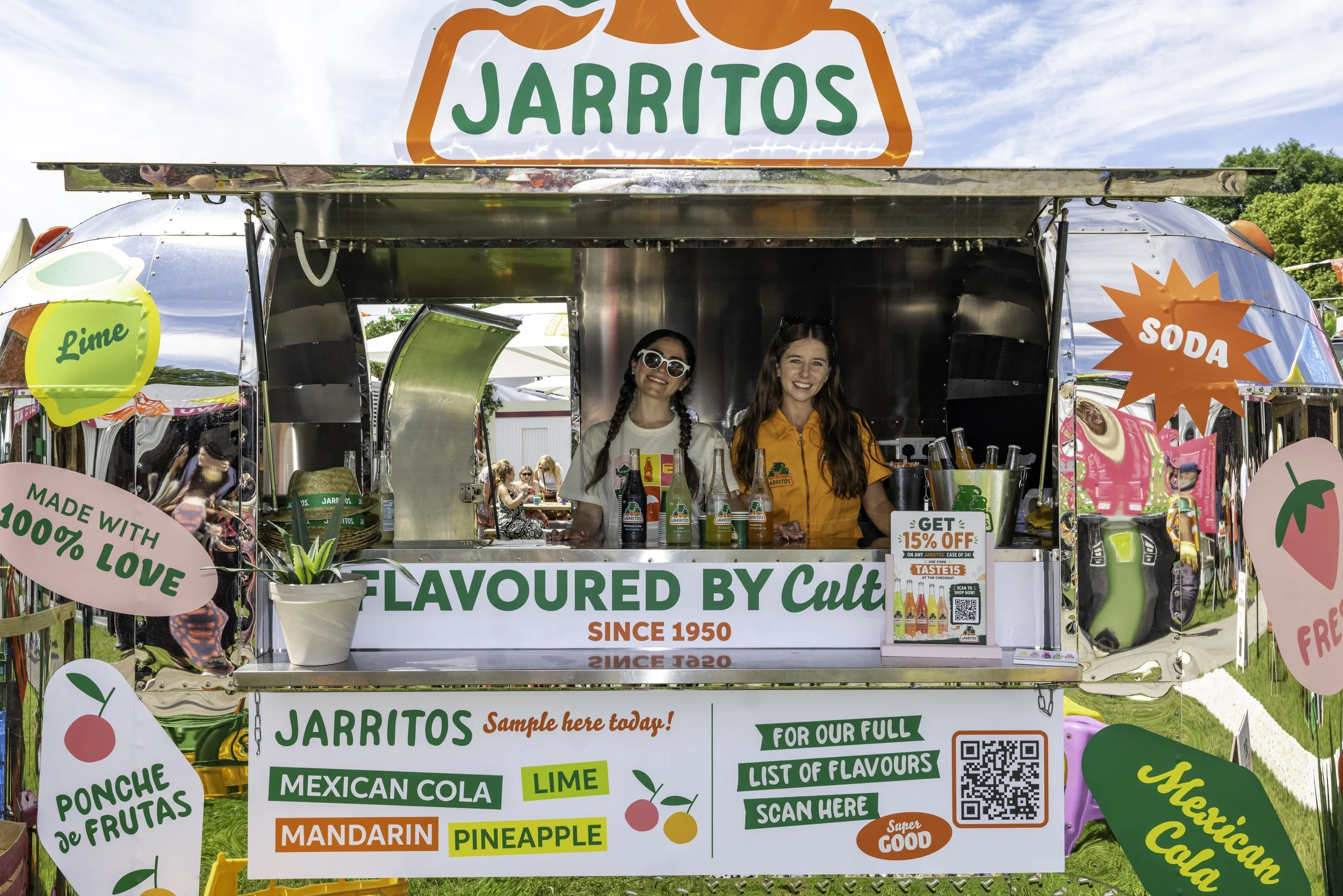 A colorful food stand for Jarritos soda with two women behind the counter smiling. The stand has signs advertising various flavors like lime, mandarin, pineapple, and more. It also mentions a 15% discount with a QR code and promotional offers.