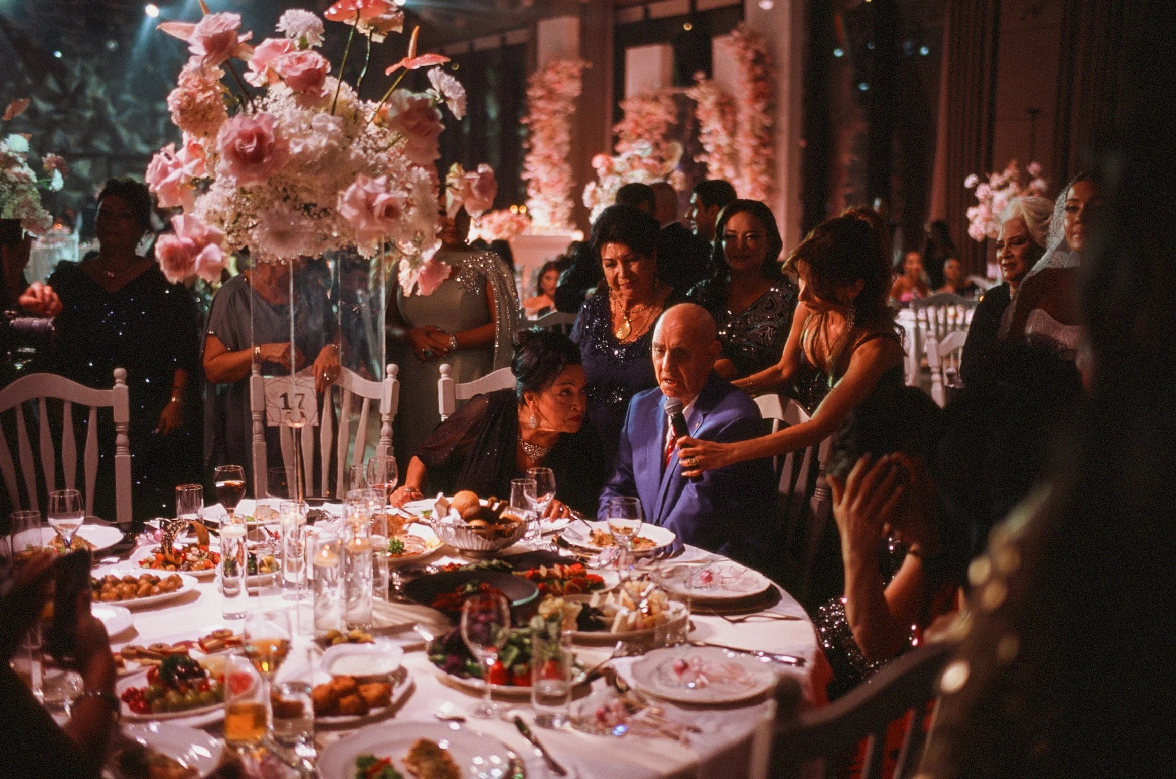 A banquet hall filled with people gathered around tables with food, flowers, and drinks, celebrating at a formal event with pink floral decorations and warm lighting.