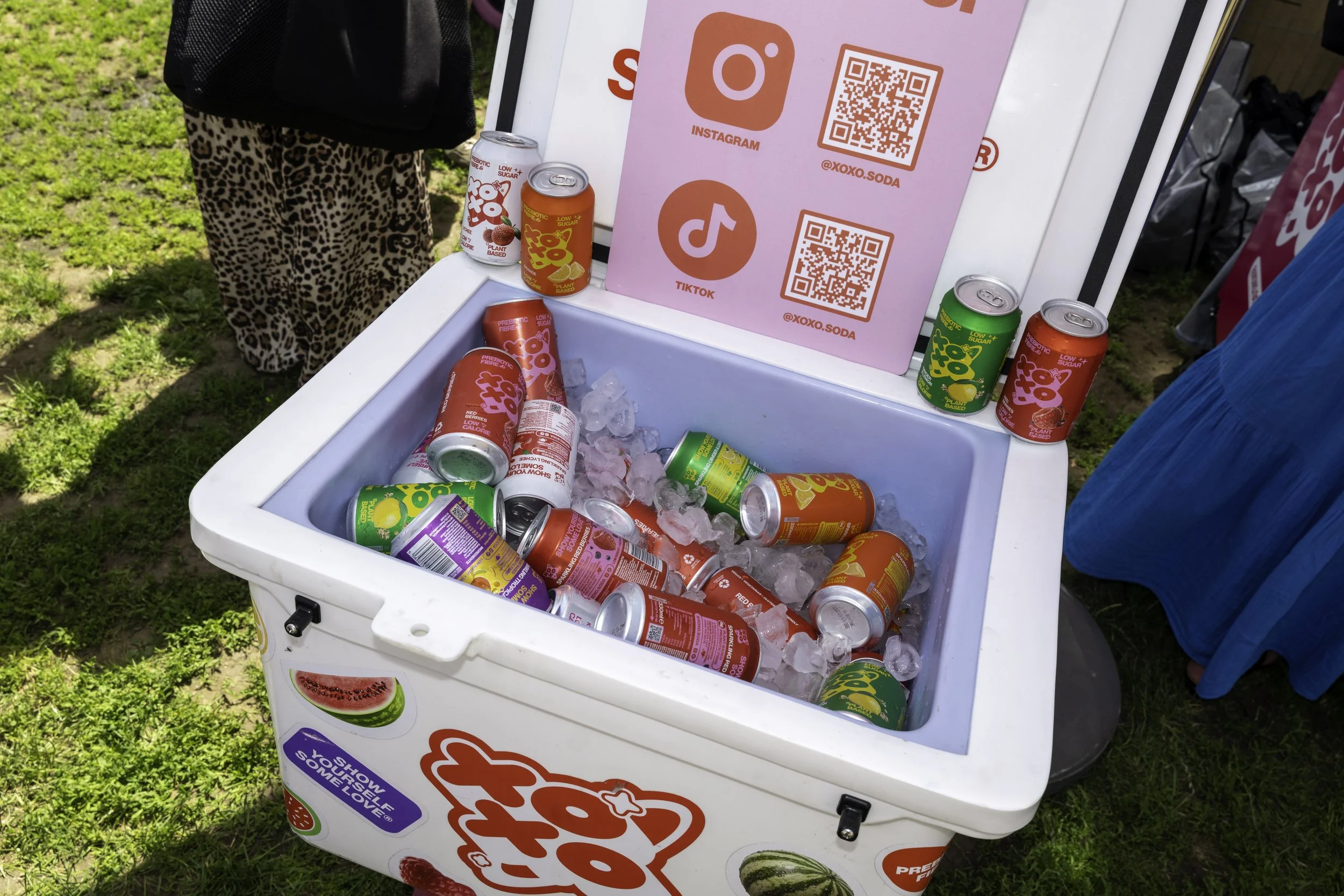 A white cooler filled with ice and various cans of soda, with a pink sign displaying QR codes and social media icons in the background. A person in leopard print pants and a black top is partially visible nearby. The cooler is outdoors on grass.