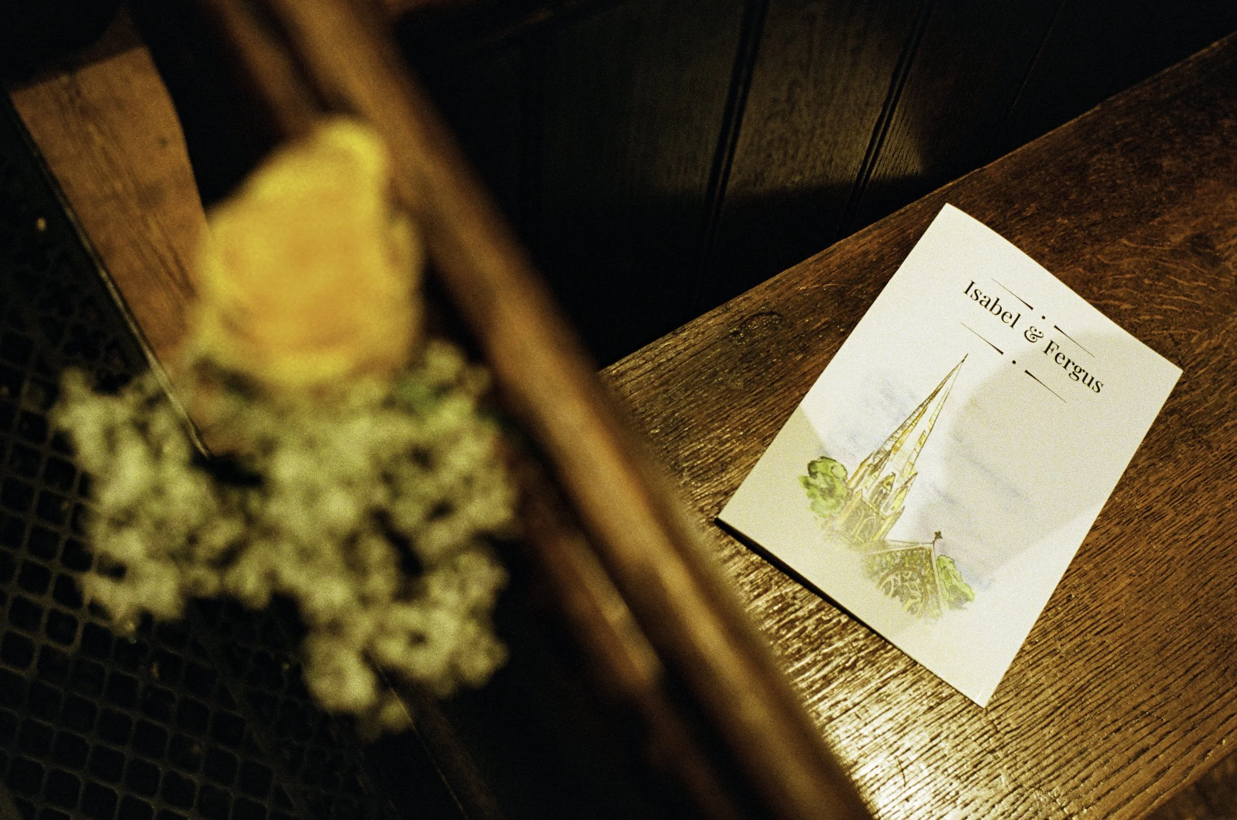 Close-up of a sushi roll with rice, seaweed, and possibly avocado or cucumber, on a black textured surface. In the background, there is a paper with the text 'Isabel & Fergus' and an illustration of a church or cathedral on a wooden table.