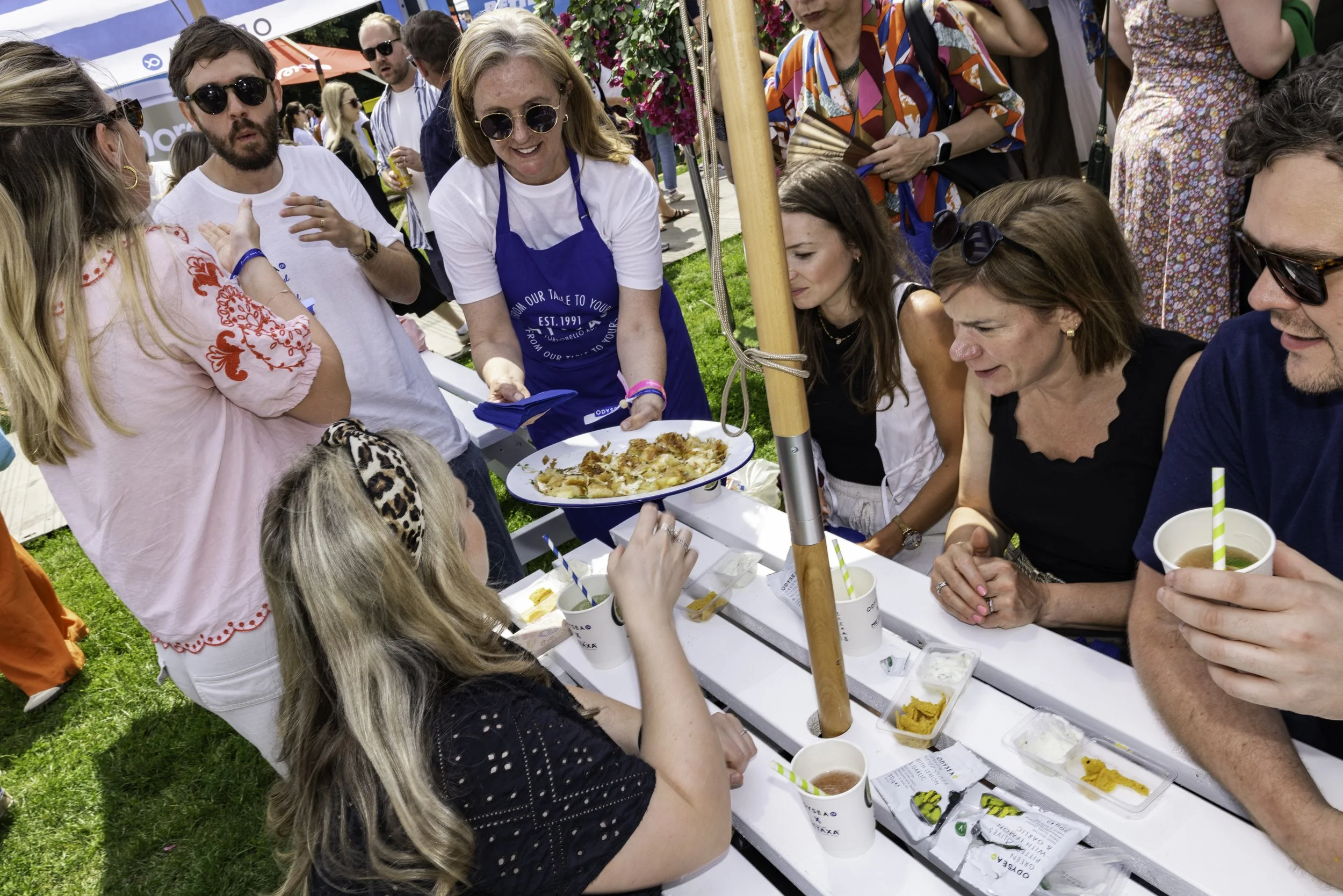 A group of people gathered around a table at an outdoor event, serving and eating pizza, with some holding drinks. There are tents and greenery in the background.