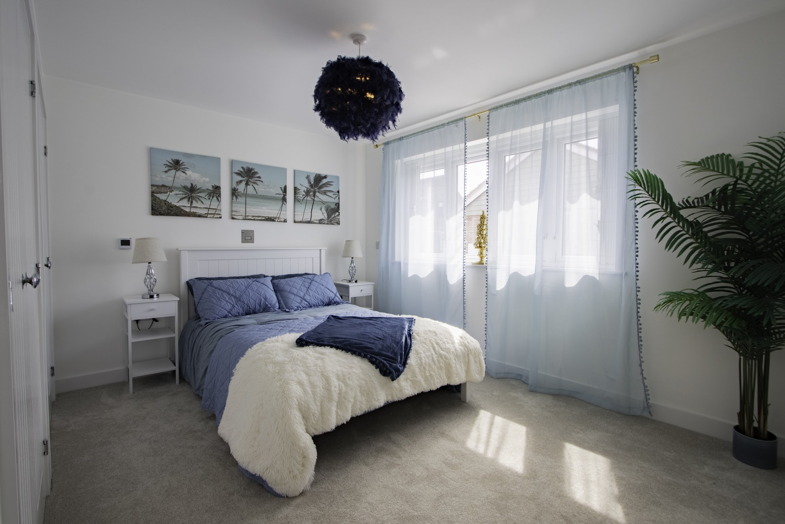 A cozy bedroom with a white bed frame, blue quilts, white nightstands with lamps, and wall art of palm trees. Light blue curtains cover a large window, and a large green potted plant is in the corner. A fluffy white blanket is draped over the bed.