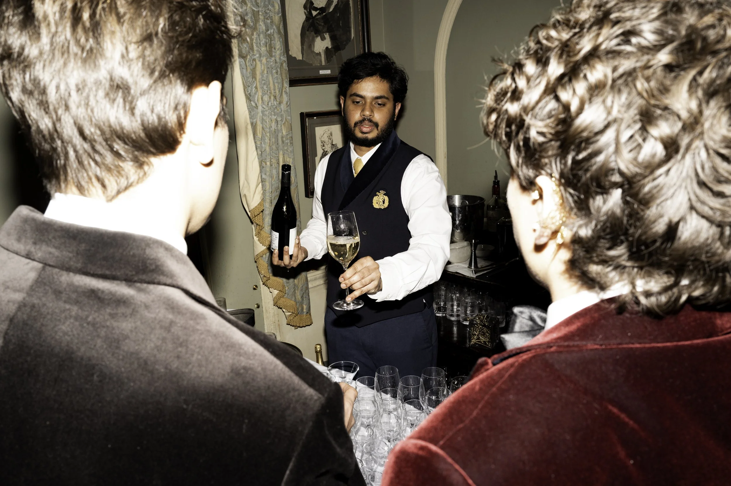 A bartender in formal attire holding a glass of white wine and a bottle, serving two women at a social gathering in a home setting.